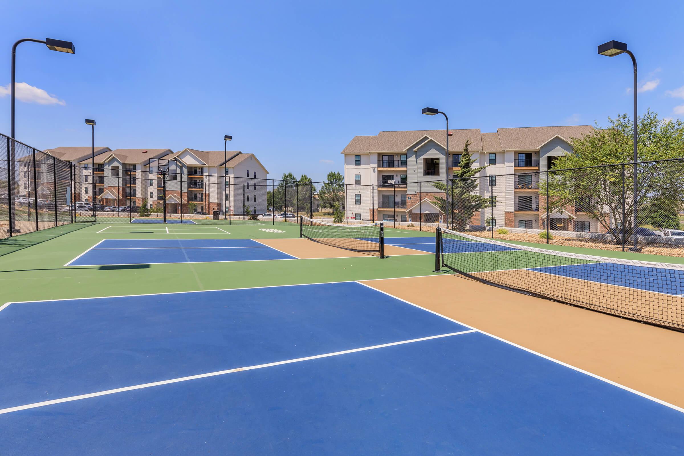 A view of multiple tennis courts featuring blue, green, and tan surfaces, surrounded by residential apartment buildings. The sky is clear and sunny, and there are light posts for illumination, suggesting a recreational area in a community setting.