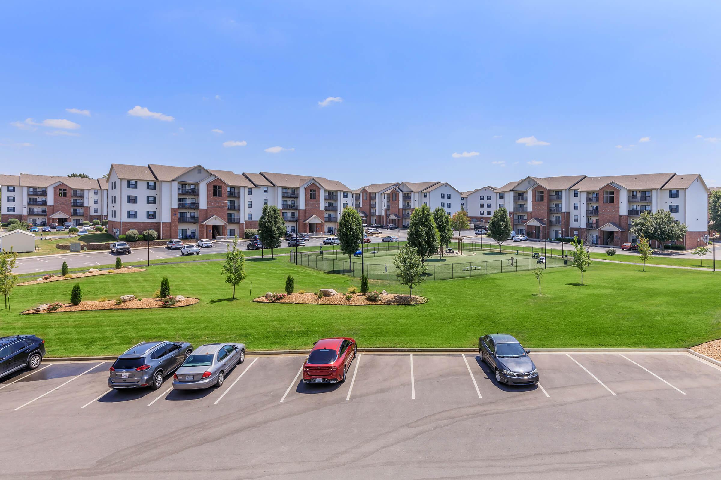 A view of an apartment complex featuring multiple buildings with a grassy area in the foreground, dotted with trees and parked cars. The sky is blue with a few clouds, and there are people playing in the open space, creating a vibrant community atmosphere.
