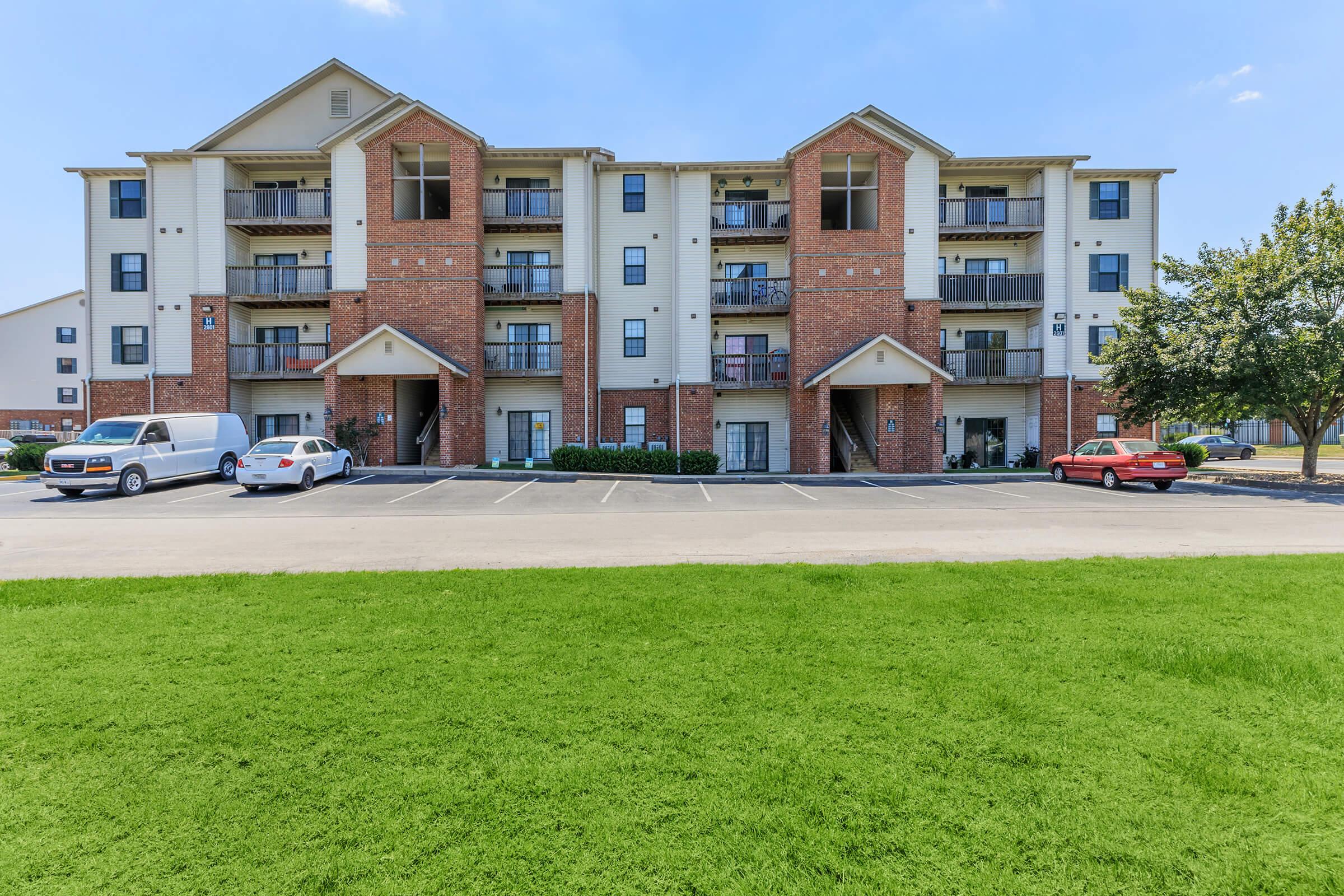A multi-unit apartment building with a red brick façade and cream-colored accents. The structure has three levels, balconies on the upper floors, and several parked cars in the foreground. A grassy area is visible in front of the building, with clear blue skies above.
