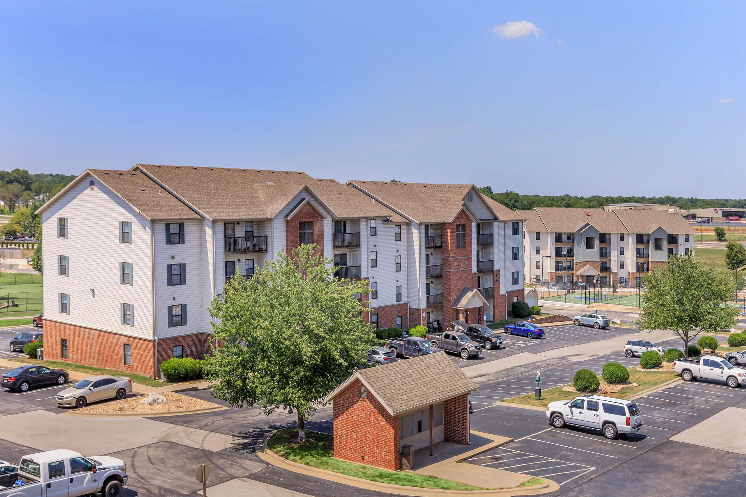 A view of a multi-unit residential apartment complex with a parking lot. The buildings are three stories high, featuring a mix of brick and siding exteriors. Nearby, there are neatly maintained grassy areas and trees, along with parked vehicles and a basketball court in the background. The sky is clear and sunny.
