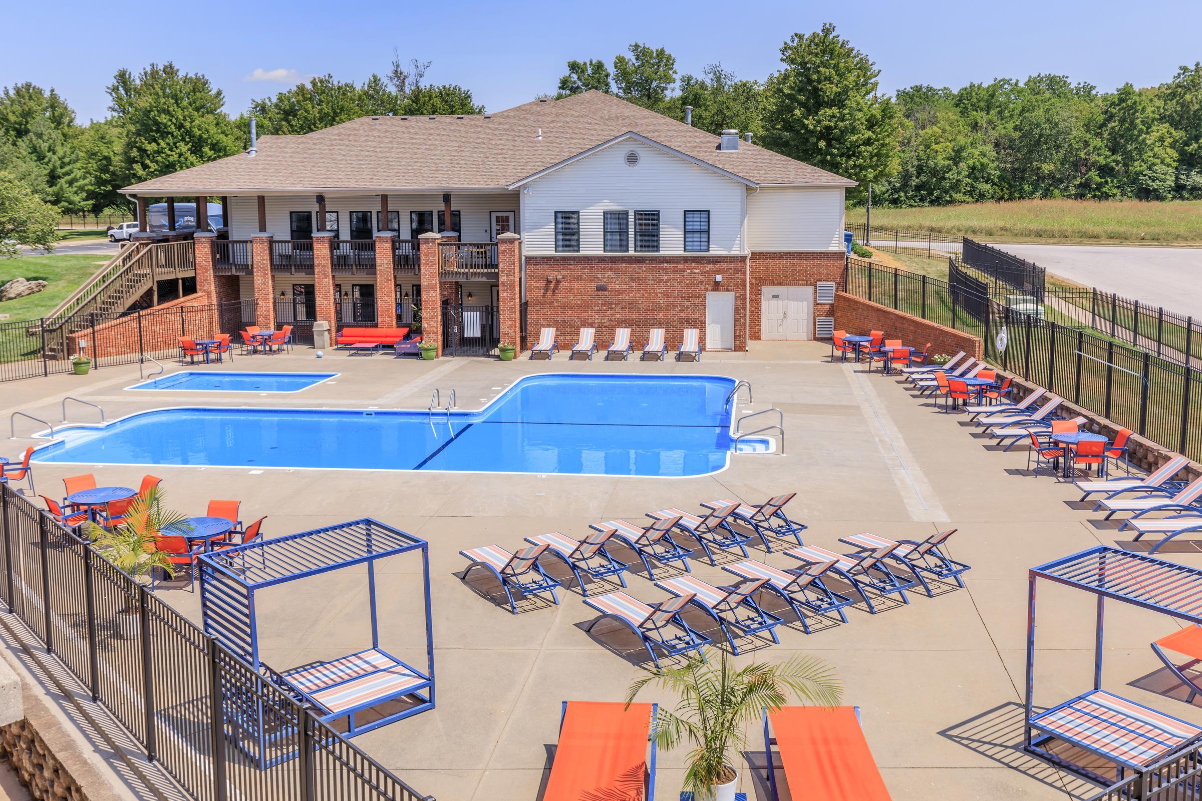 A spacious outdoor swimming pool area with lounge chairs and vibrant red seating. The pool is surrounded by a safety fence, and a building featuring brick and white siding is in the background. Lush greenery and trees are visible around the area under a clear blue sky.