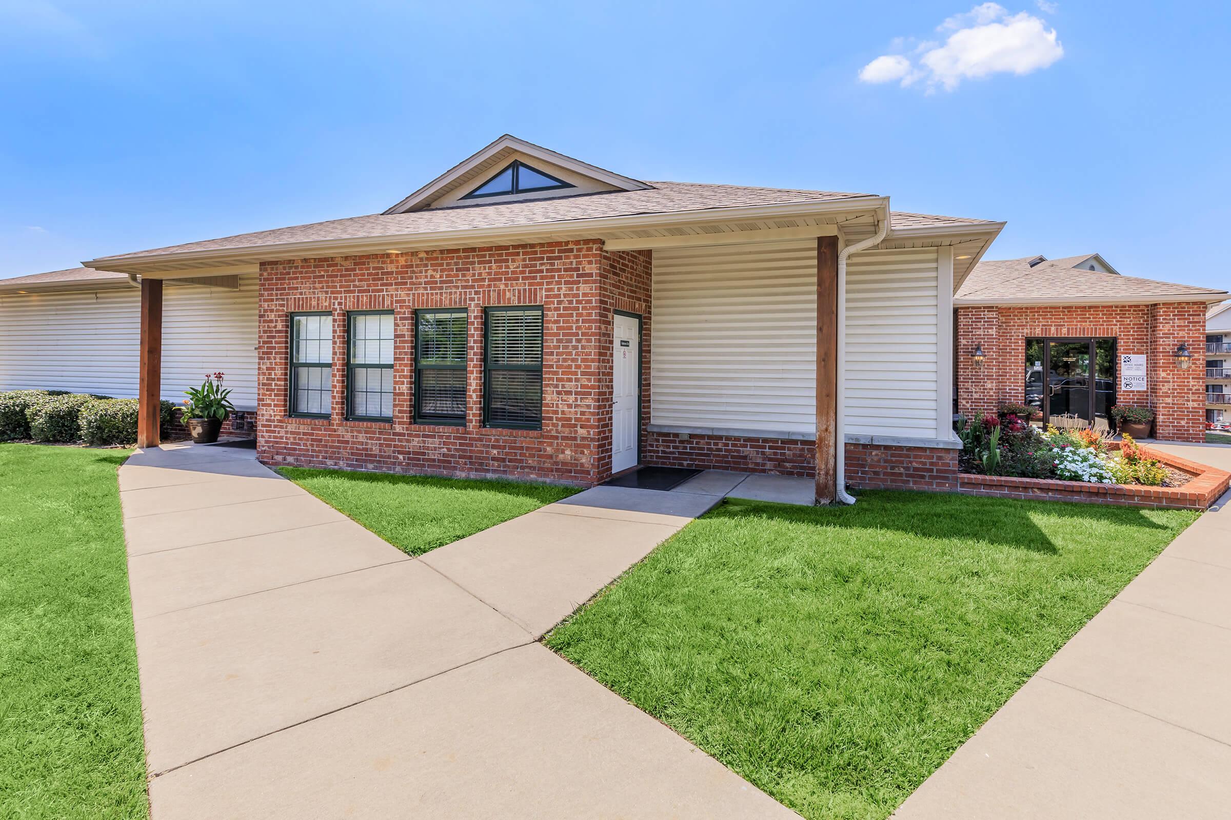 A single-story brick building with a light-colored wooden exterior. Large windows are seen on the front, and a pathway leads to the entrance. Lush green grass and decorative plants surround the area, with a clear blue sky overhead.