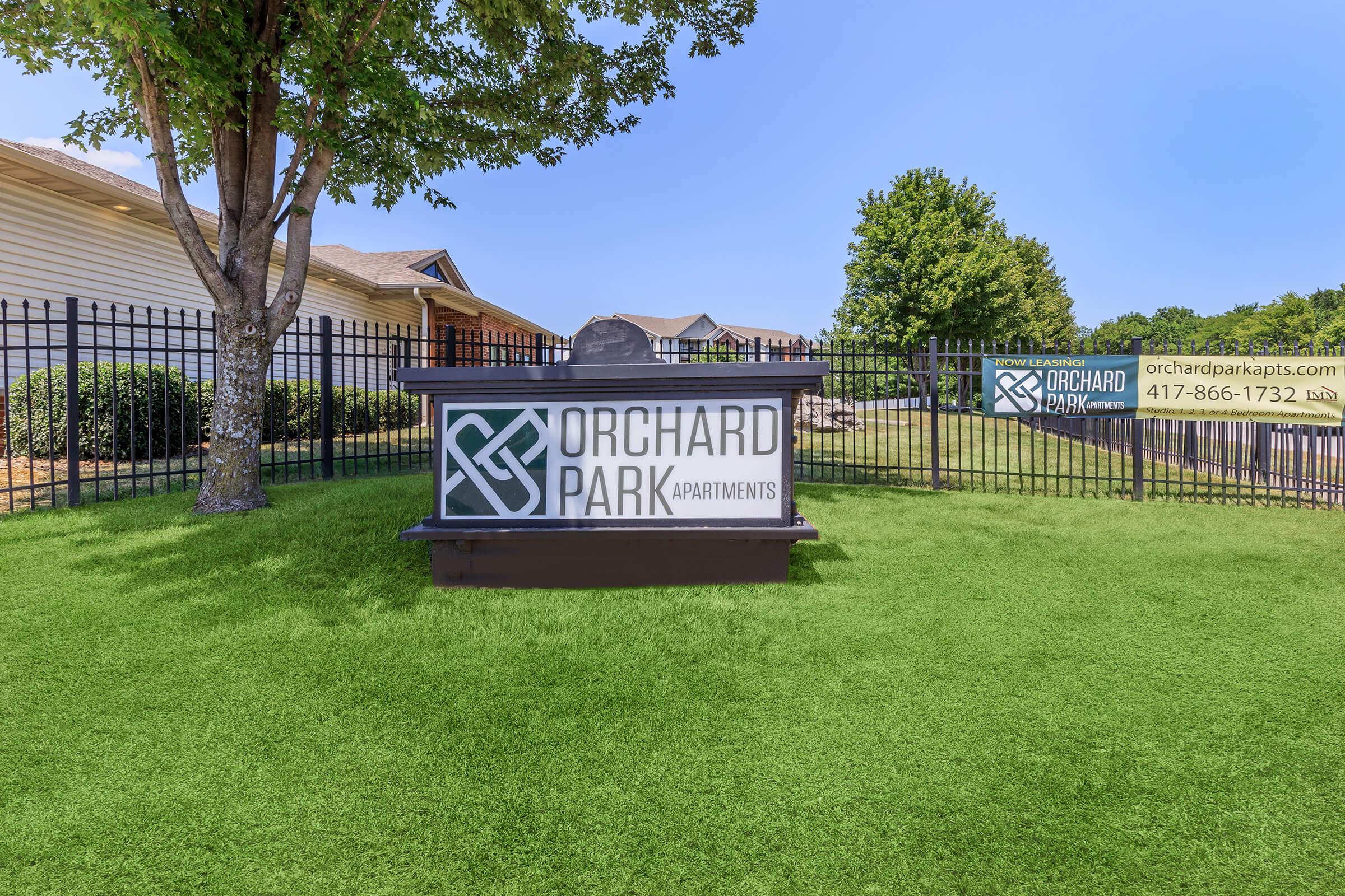 A sign for Orchard Park Apartments displayed in front of a landscaped area with green grass, trees, and a blue sky in the background. The sign features the name "Orchard Park" and includes contact information and a website for the apartments.