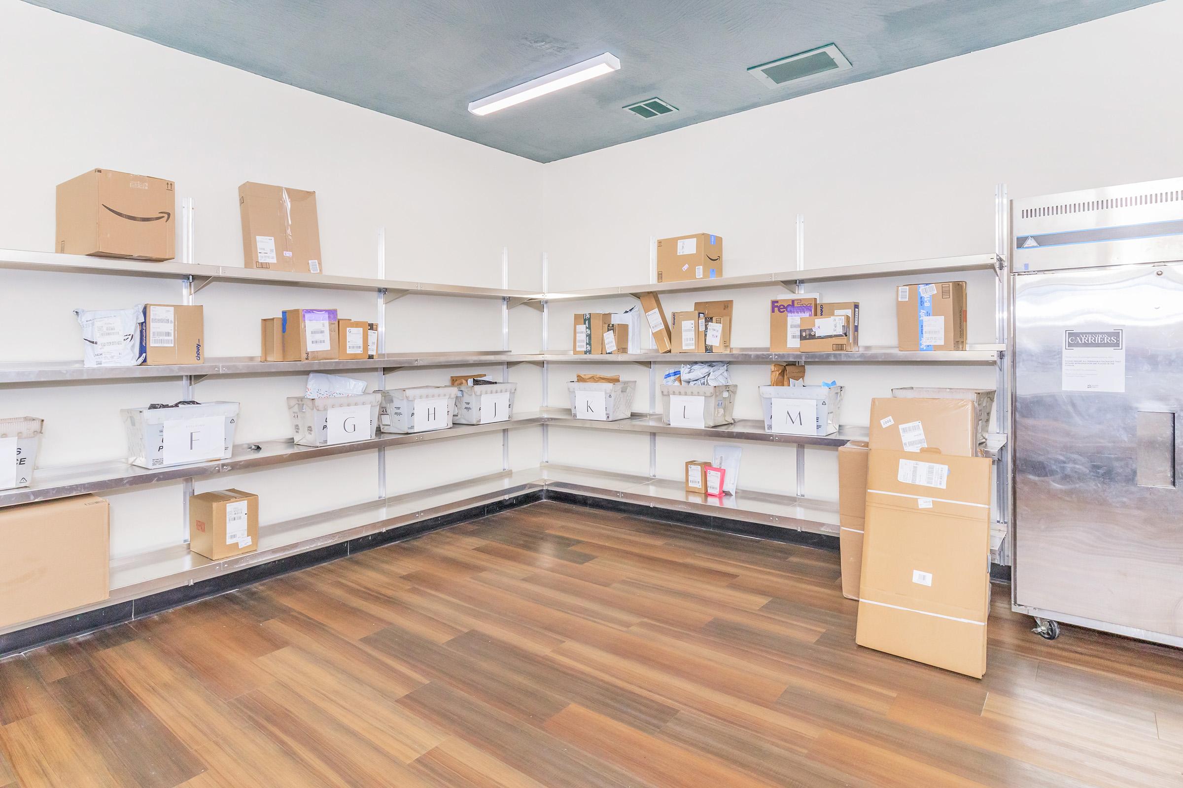 An empty storage room with wooden flooring and white walls. There are several shelves filled with cardboard boxes of various sizes, some labeled with shipping information. A stainless steel refrigerator is visible in the corner, and the overall space appears clean and organized.