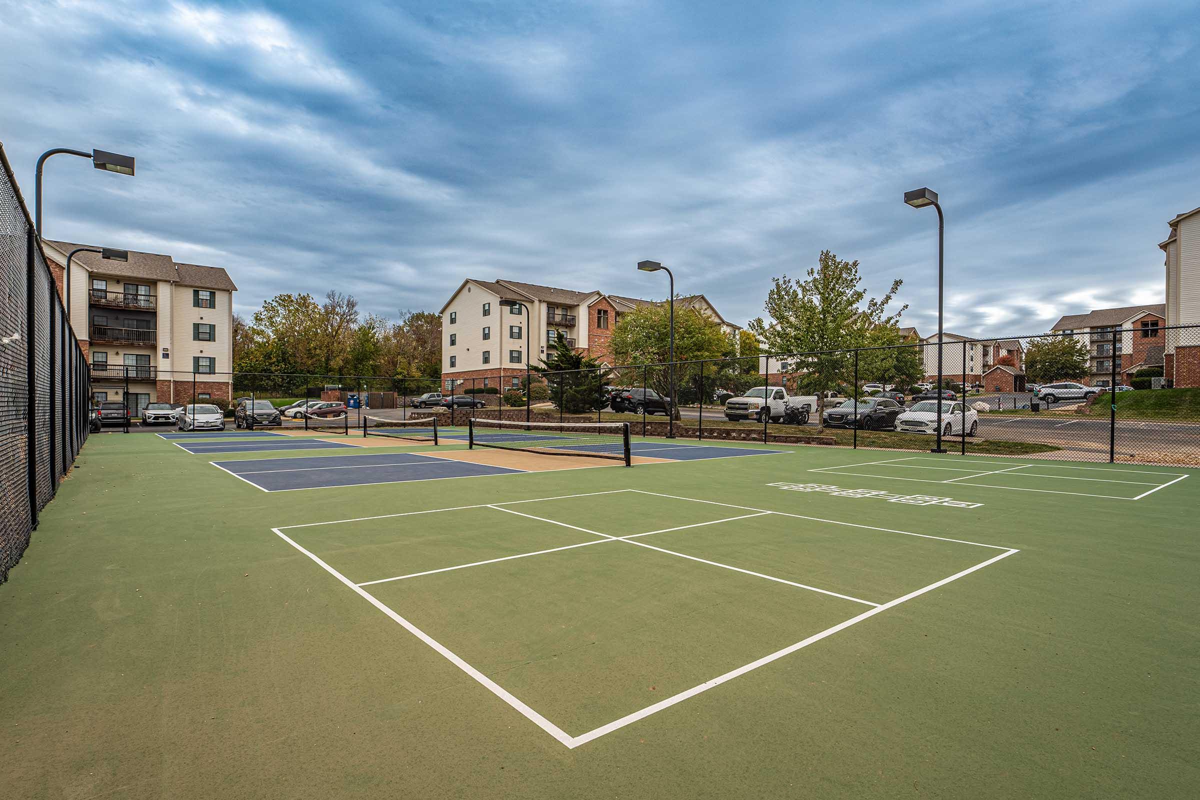 View of a tennis court with green and brown surfaces, surrounded by residential buildings and parked cars. The sky is partly cloudy with a mix of gray and blue. There are several lights on poles illuminating the court area.