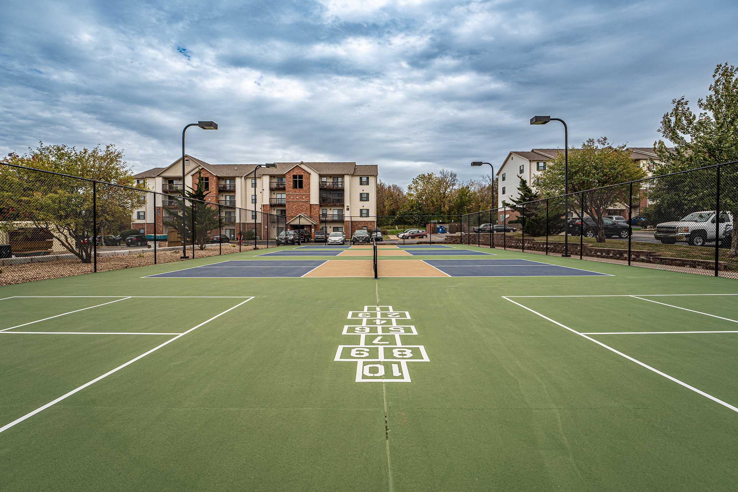 A view of a multi-use sports court featuring hopscotch markings and divided playing areas, surrounded by an apartment complex and landscaping. The sky is overcast with gray clouds, and parked cars are visible in the background.
