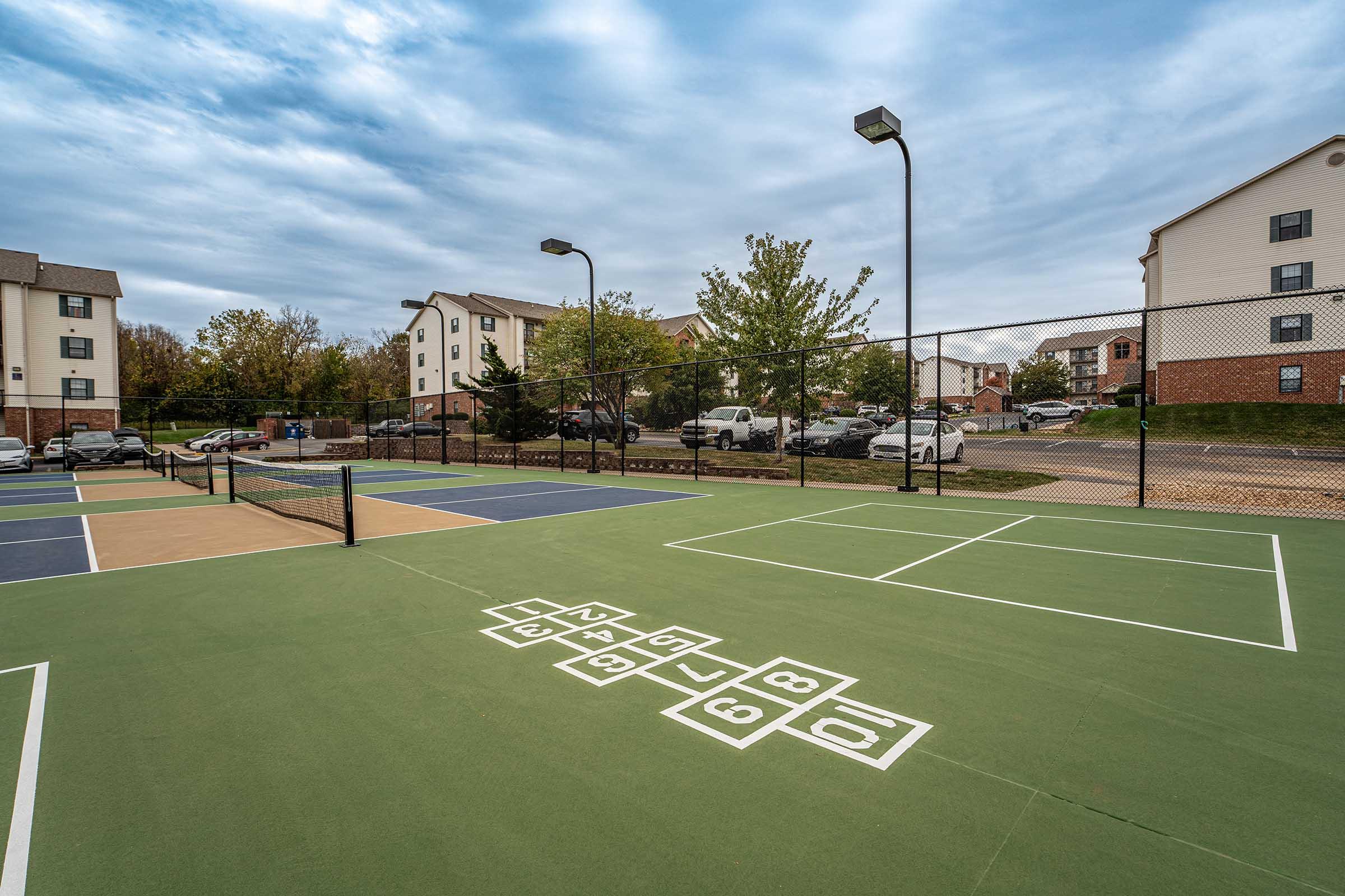 A view of a tennis court with green and blue surfaces in an outdoor recreational area. In the foreground, there's a hopscotch design on the court. Behind the courts, there are residential buildings and parked cars under a cloudy sky.