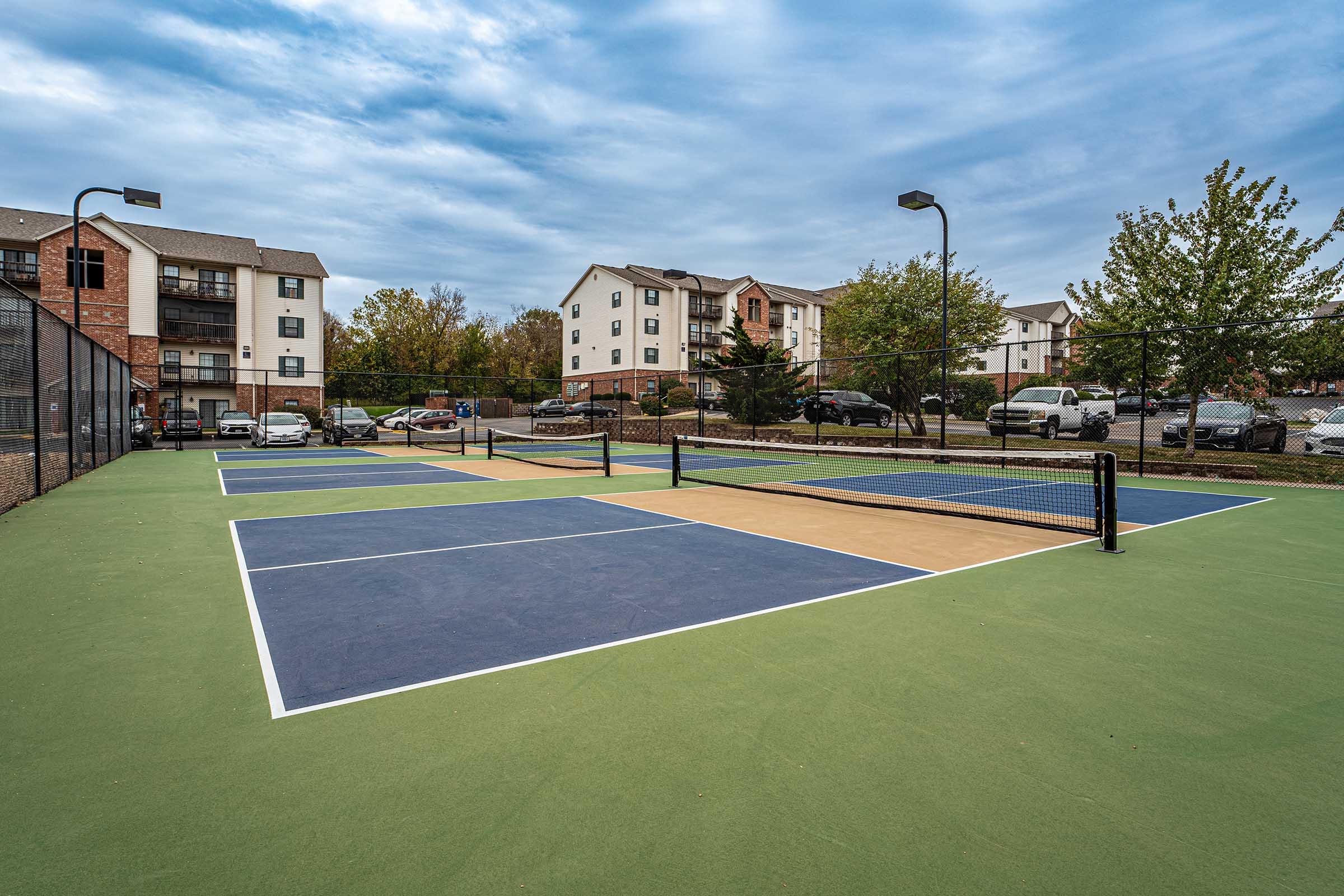 Newly constructed tennis courts with blue and tan surfaces, surrounded by green grass. In the background, several apartment buildings and a parking lot with cars are visible under a cloudy sky.