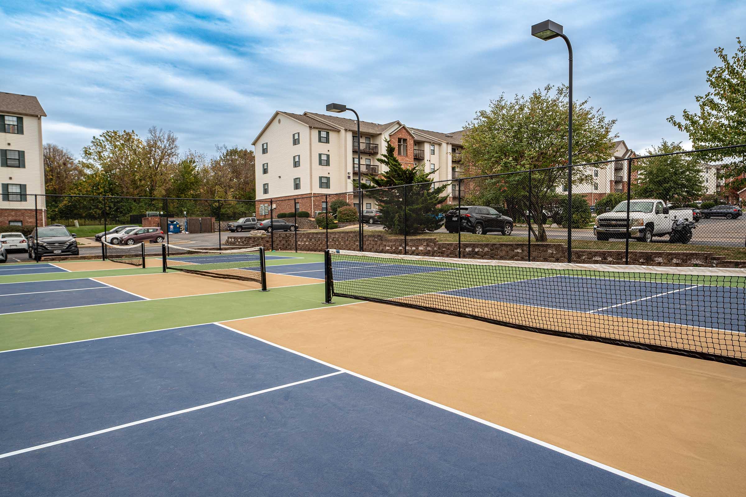A view of outdoor tennis courts featuring multiple colored surfaces—blue, green, and beige. In the background, there are residential buildings surrounded by trees and parked cars. The sky is overcast, suggesting a cloudy day.