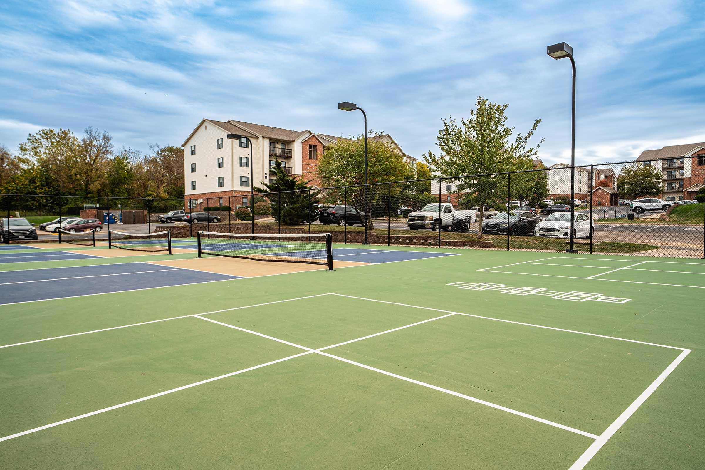 View of a vacant outdoor tennis court with green and blue playing surfaces, surrounded by trees and residential buildings in the background. Several parked cars are visible nearby, and the sky is overcast.
