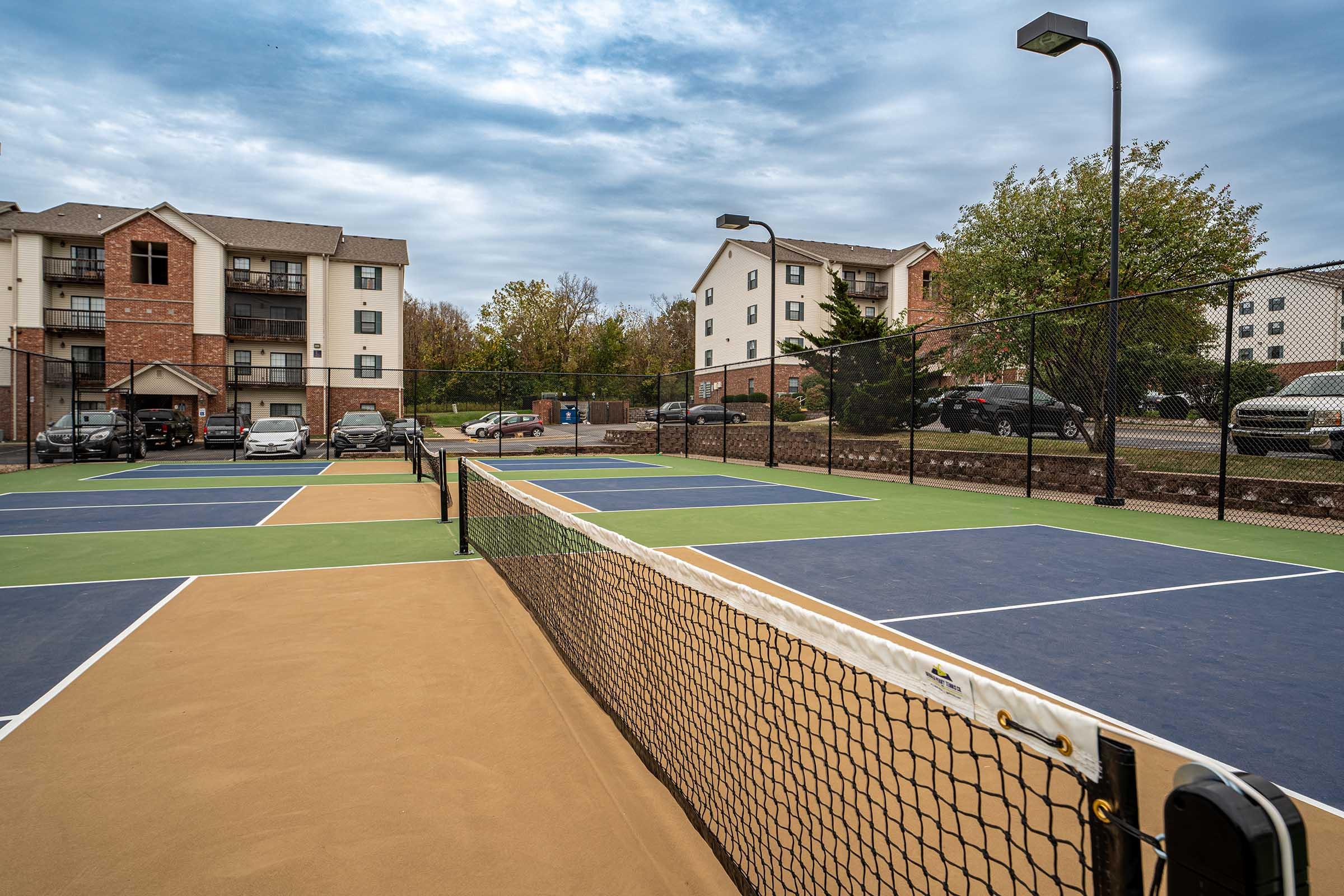 View of a tennis court with a net dividing it in the foreground. The court features blue and tan surfaces. In the background, there are residential buildings and parked cars. The sky is overcast, suggesting a cloudy day.