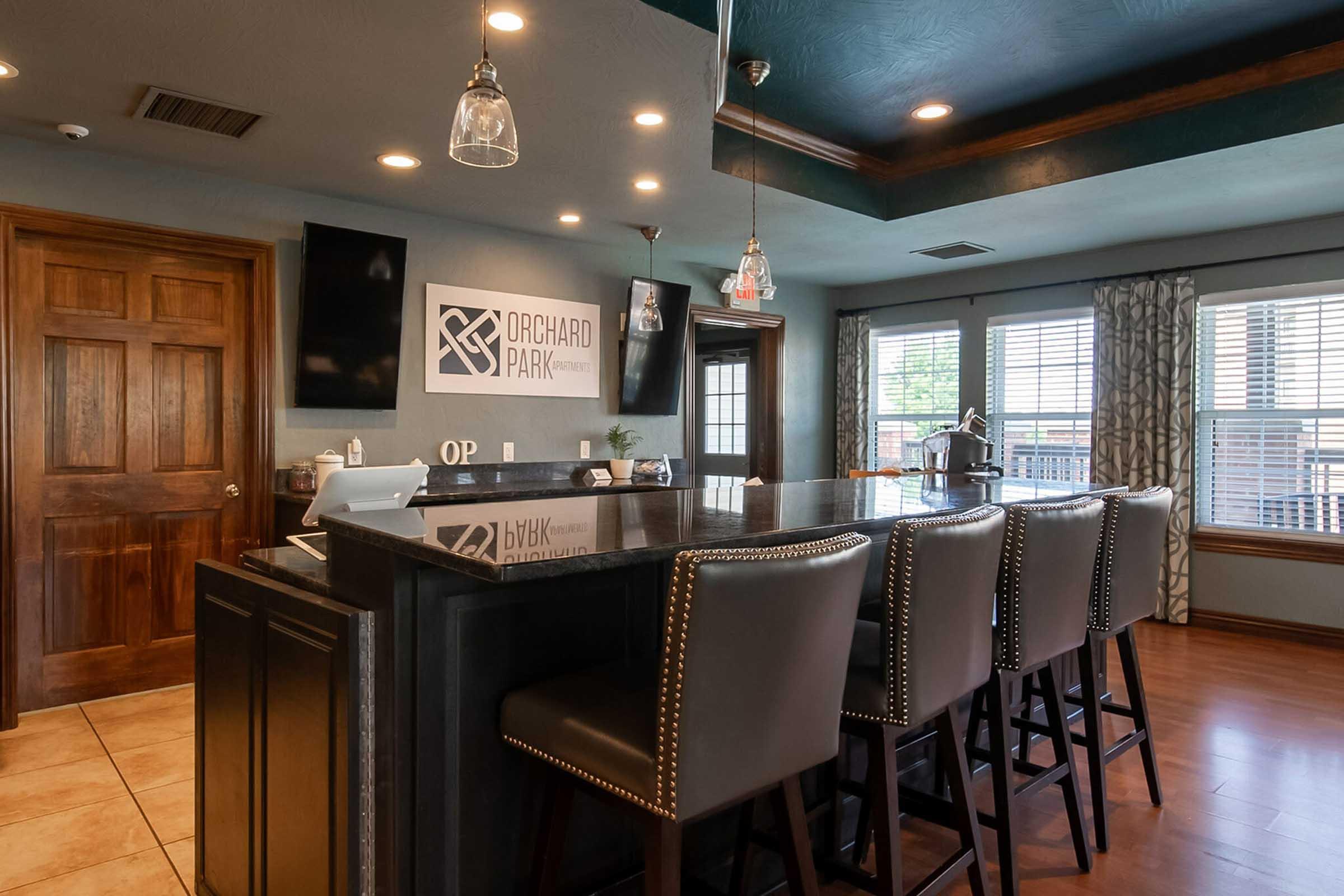 A stylish interior of a bar area featuring dark wood accents, a black countertop, and several high-backed chairs. A logo for "Orchard Park" is visible on the wall, along with large windows letting in natural light and decorative curtains. The space has a modern yet cozy atmosphere.