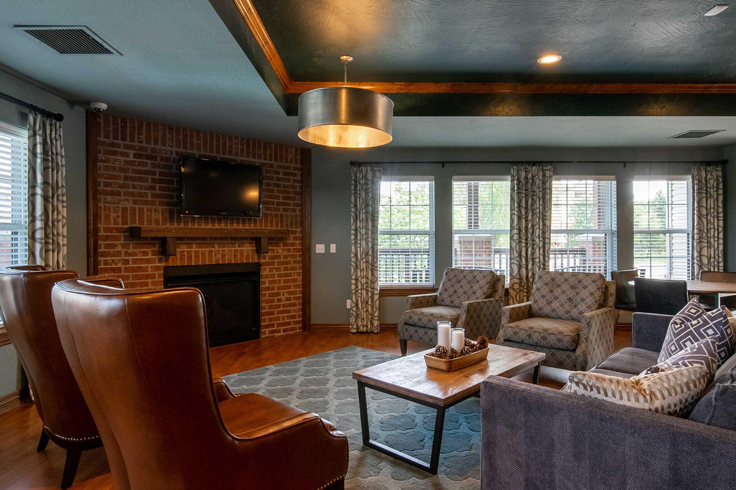 Cozy living area featuring a mix of modern and rustic design elements. There are two stylish lounge chairs, a sofa, and a wooden coffee table adorned with decorative items. The room has a stone fireplace, a wall-mounted TV, large windows letting in natural light, and a patterned area rug on hardwood floors.
