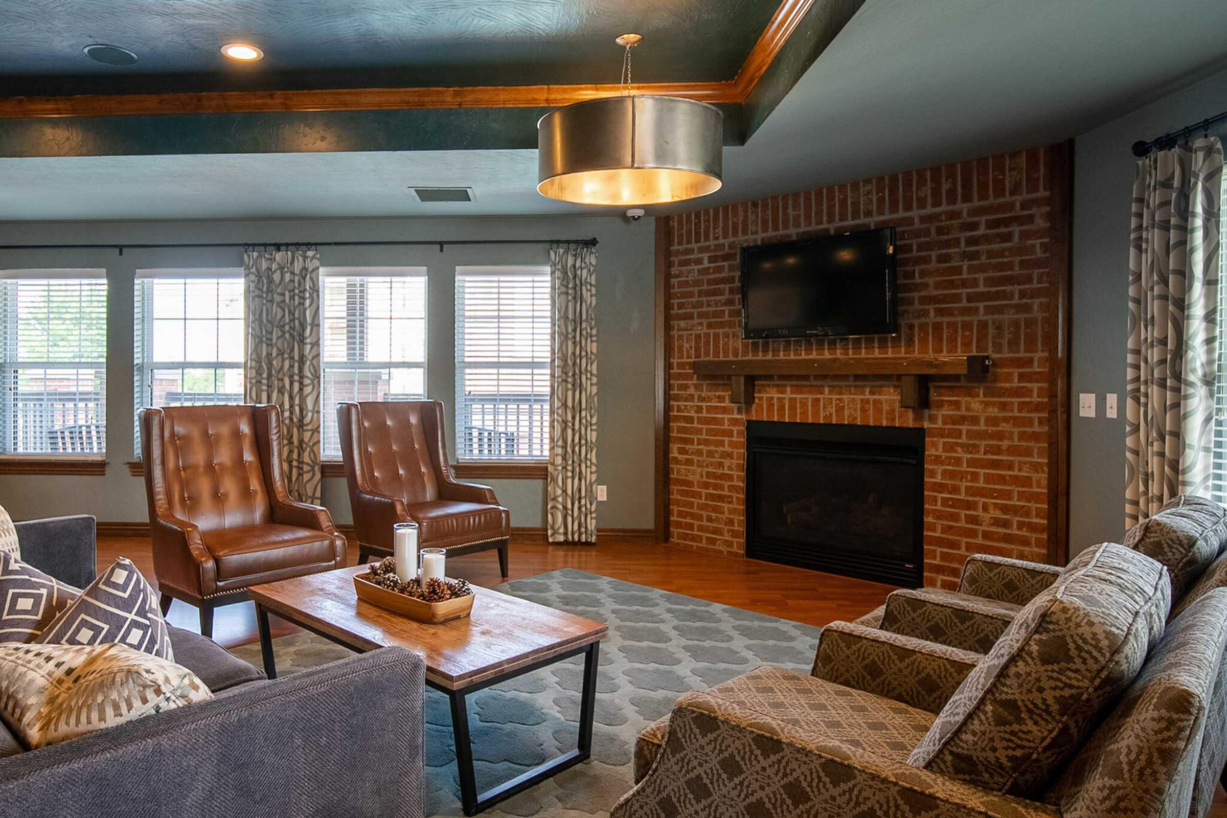 A cozy living room featuring two brown armchairs, a comfortable gray sofa, and a wooden coffee table. The room has large windows with patterned curtains, a brick fireplace, and a wall-mounted TV, creating a warm and inviting atmosphere. A decorative centerpiece is placed on the coffee table.