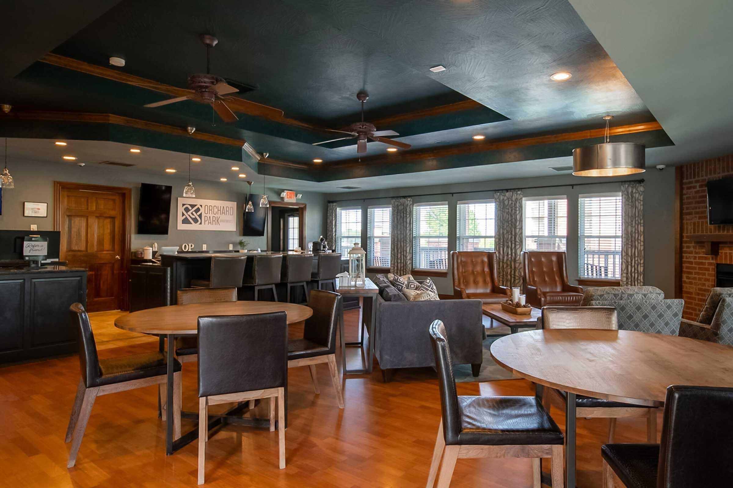 A modern common area featuring round and rectangular wooden tables, black leather chairs, and comfortable seating. Large windows allow natural light, and fans hang from the ceiling. The décor includes dark wood accents and stylish light fixtures, creating a welcoming atmosphere for gatherings or relaxation.