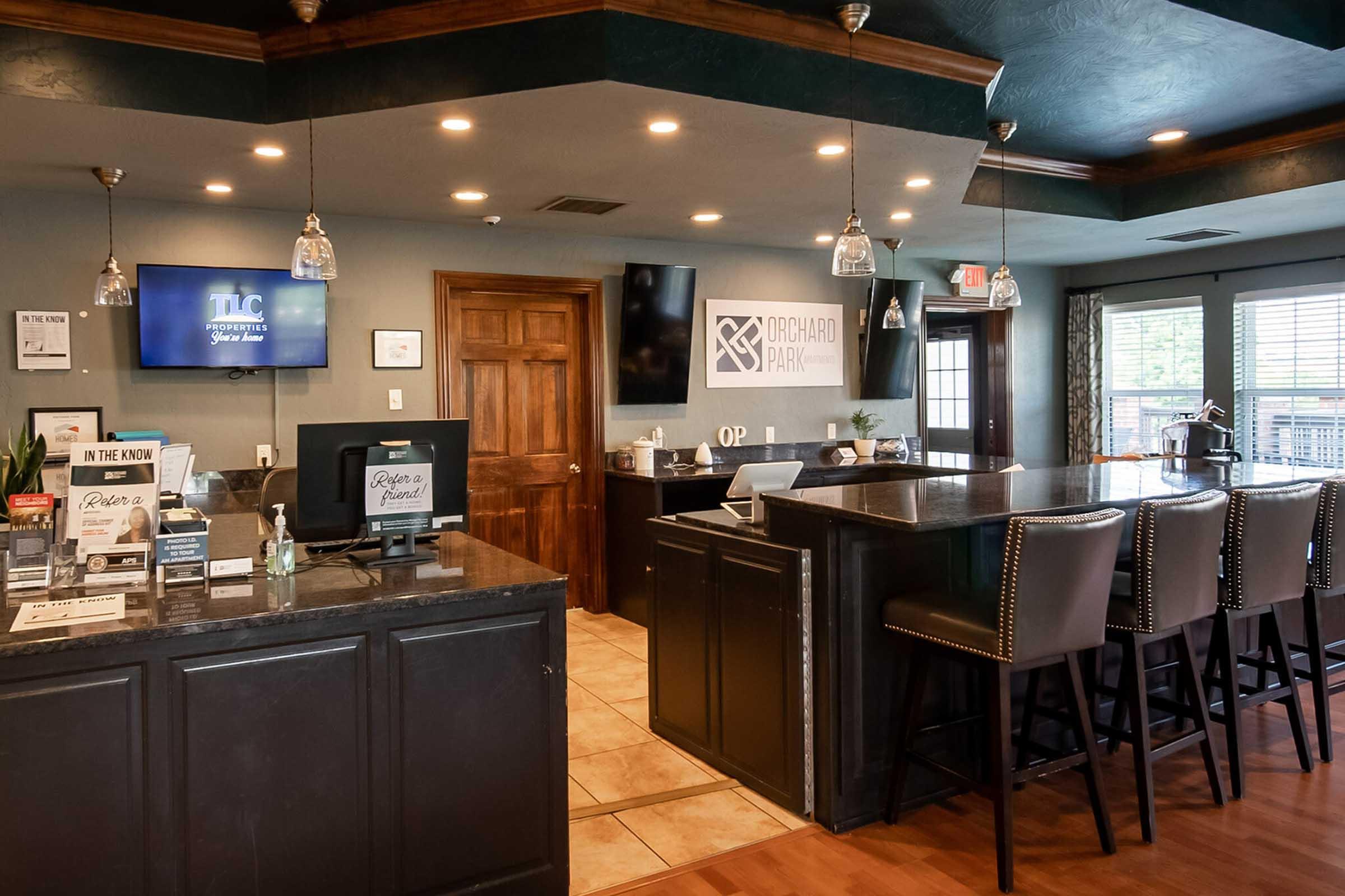 Interior view of a modern reception area featuring a dark countertop with a cash register, bar stools, and promotional materials on display. There are light fixtures hanging from the ceiling and a logo on the wall. Large windows provide natural light, and the space has a warm, inviting atmosphere.