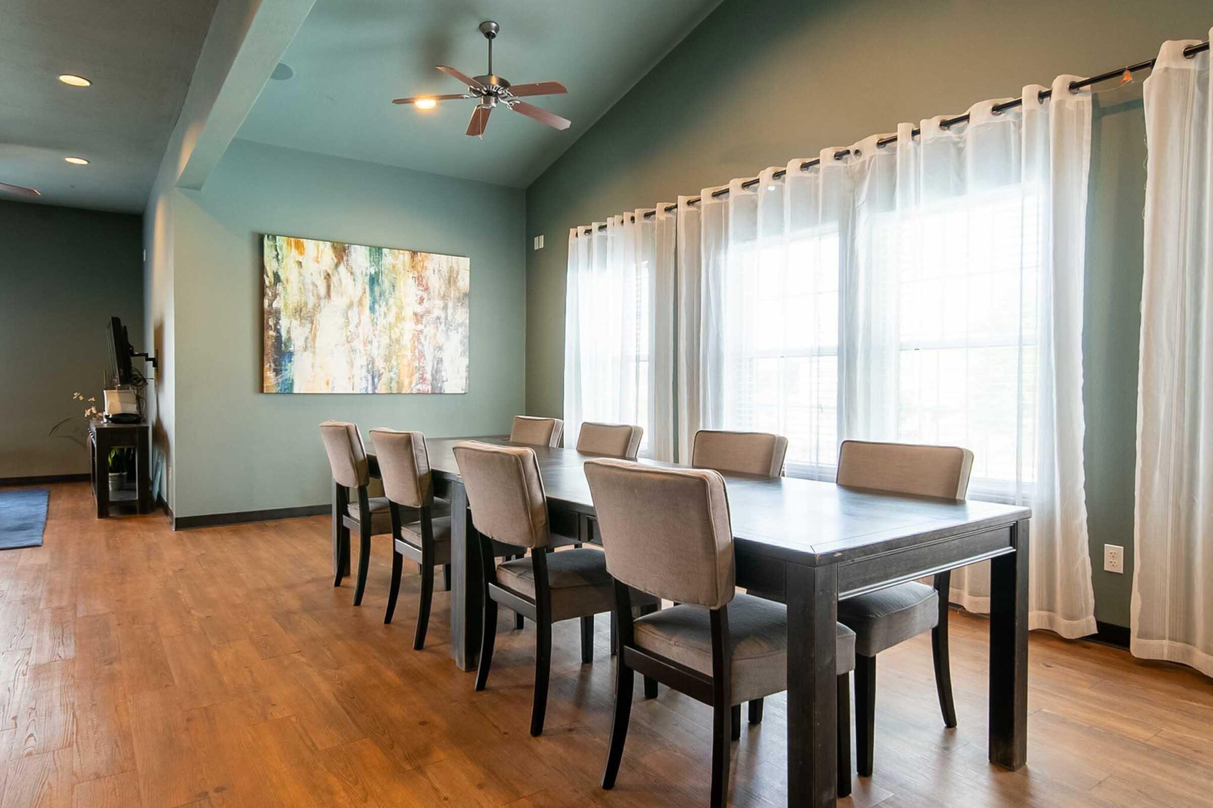 A modern dining area featuring a wooden table with eight upholstered chairs, set against a backdrop of light green walls and large windows covered with sheer white curtains. An abstract painting hangs on the wall, adding a pop of color to the space. The floor is made of light hardwood.