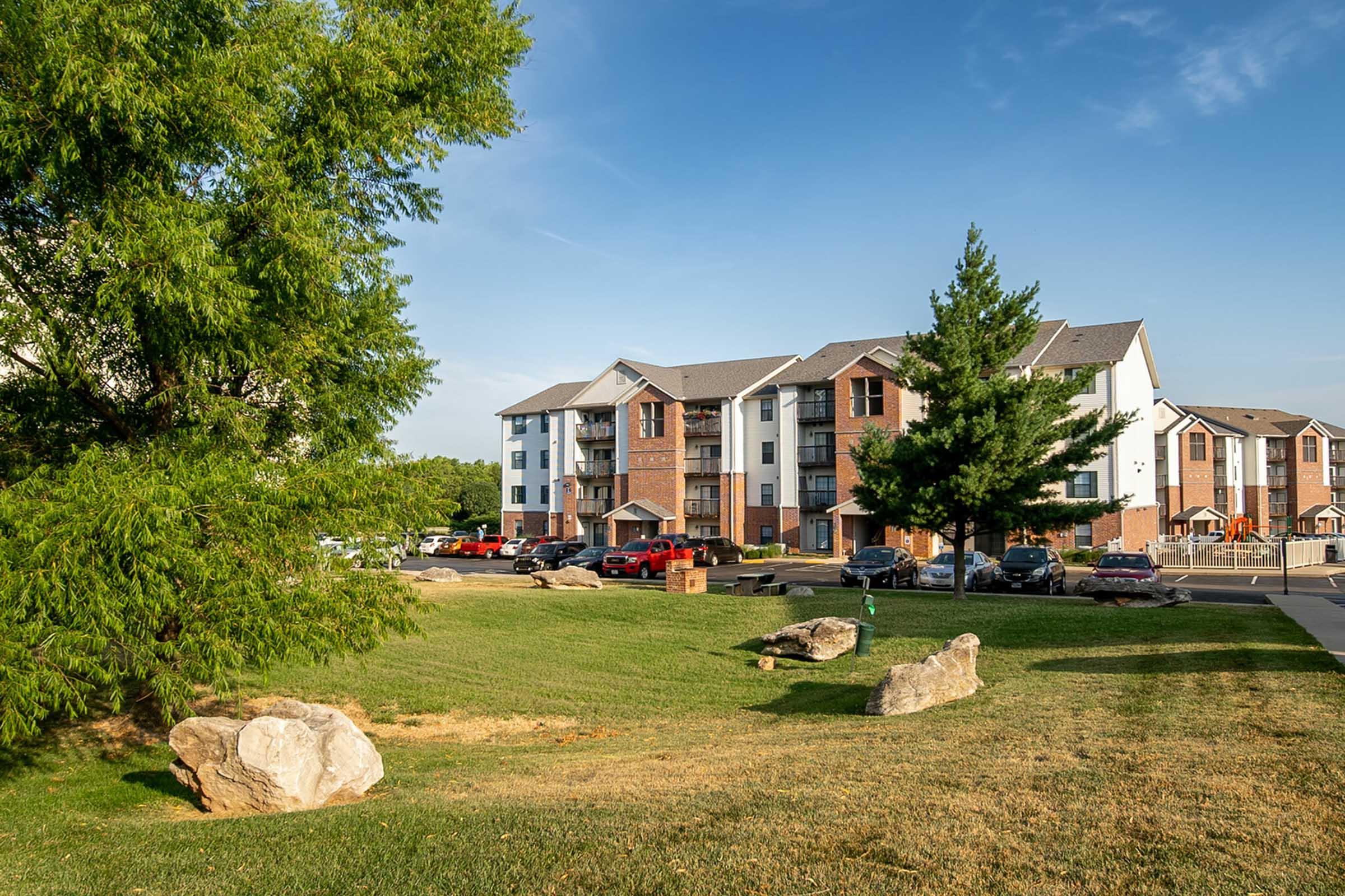 A view of a residential apartment complex featuring three-story buildings in a suburban setting. The foreground includes a grassy area with large rocks and trees, while parked cars are visible in the lot. The sky is clear with some clouds, suggesting a pleasant day.