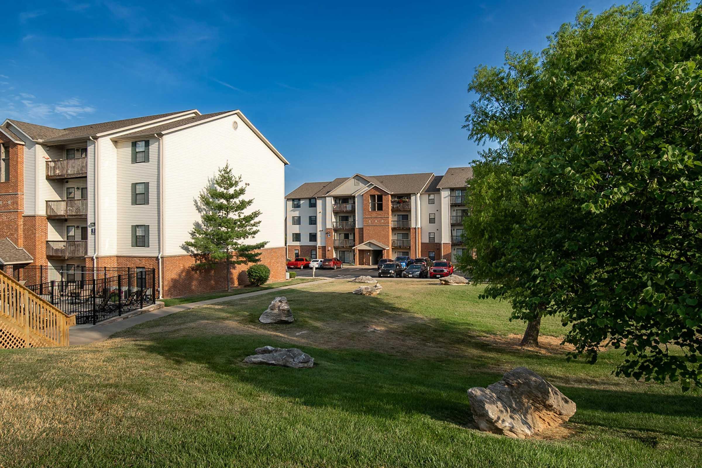 A view of a residential complex featuring multiple apartment buildings and a green lawn. Large rocks are scattered on the grass, and there are trees providing shade. The sky is clear and blue, indicating a sunny day. Vehicles are parked in front of the buildings, creating an inviting atmosphere.