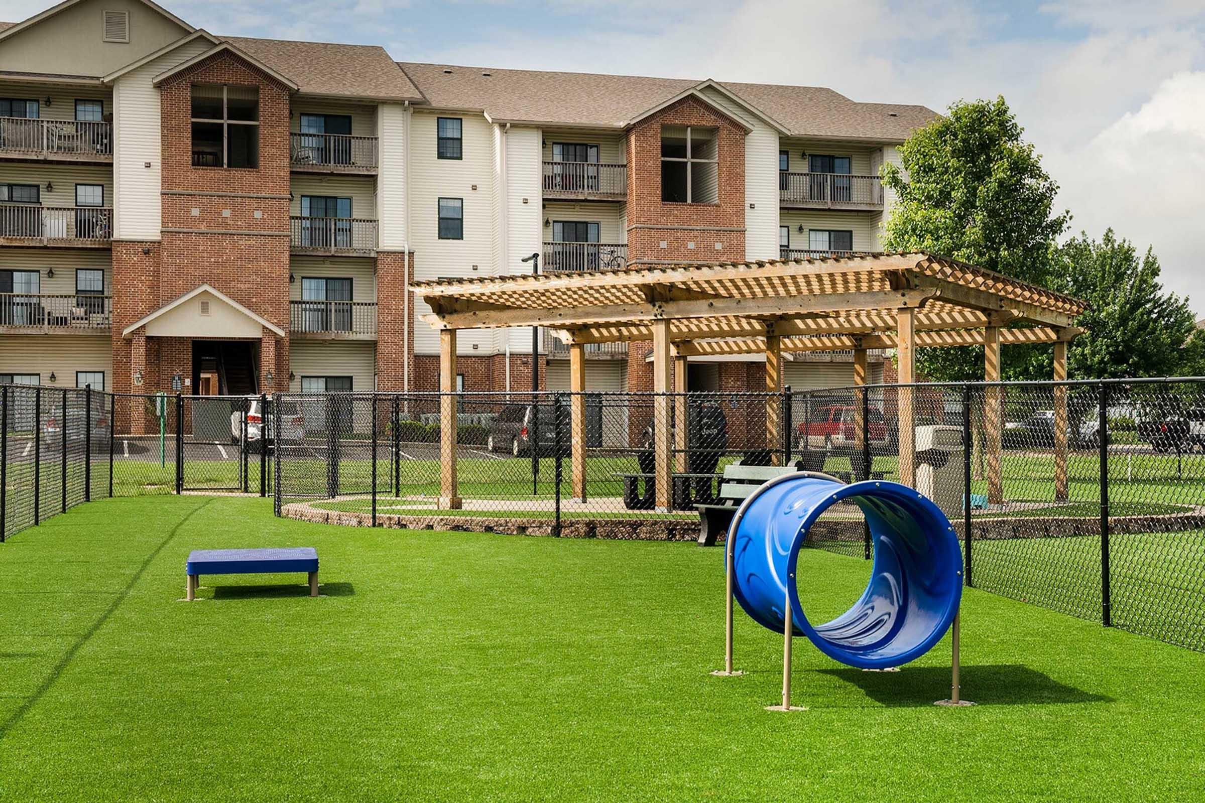 Playground area featuring a blue tube slide and a bench on green artificial grass, surrounded by a black fence. In the background, there are multi-story apartment buildings with balconies. A wooden pergola provides shade near the playground. The sky is partly cloudy.