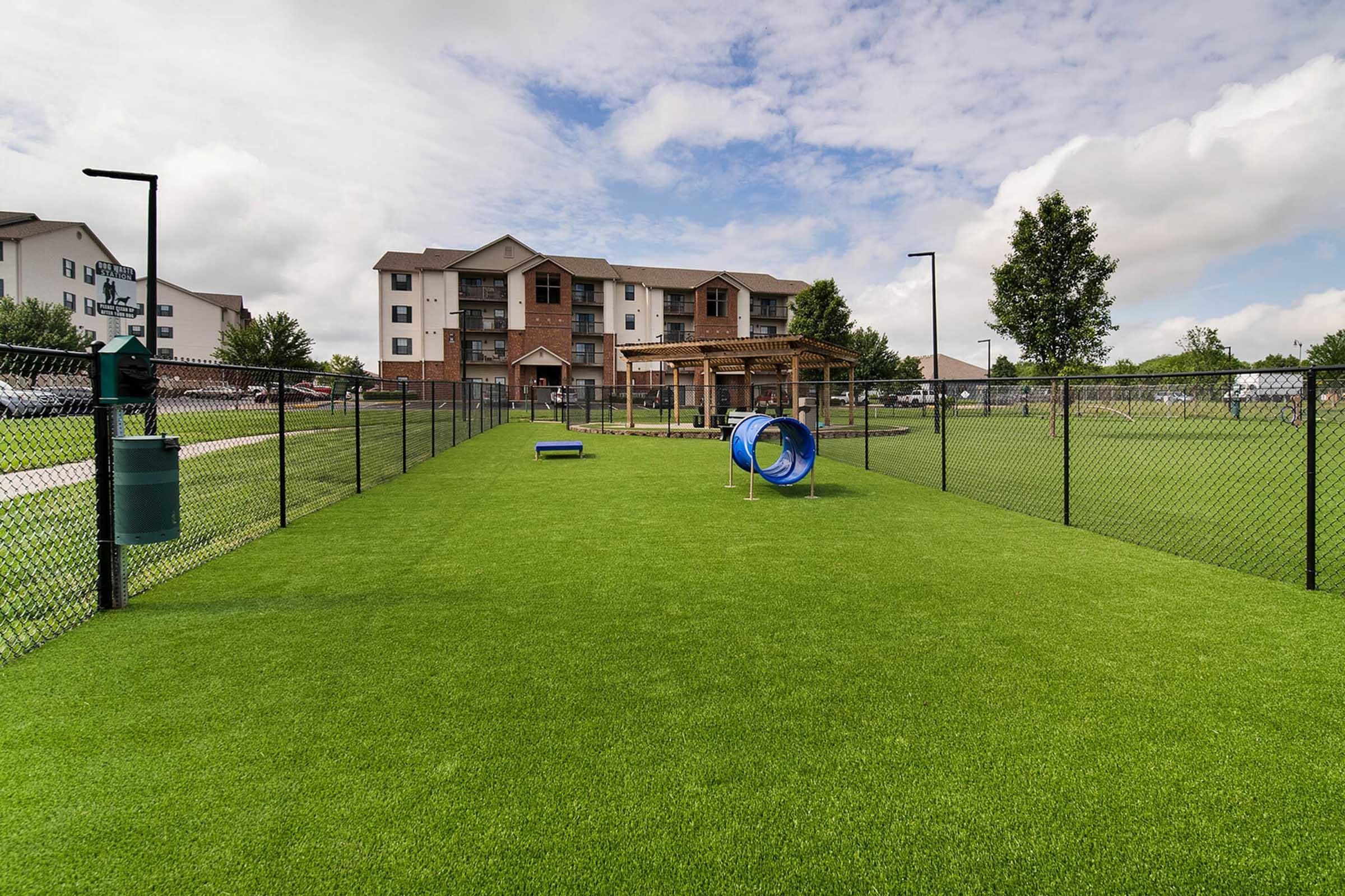 A spacious dog park featuring artificial grass, a blue tunnel, and a small agility obstacle. In the background, a multi-story residential building is visible, set against a partly cloudy sky. The area is enclosed with a black fence, and there are trees and pathways around the park.
