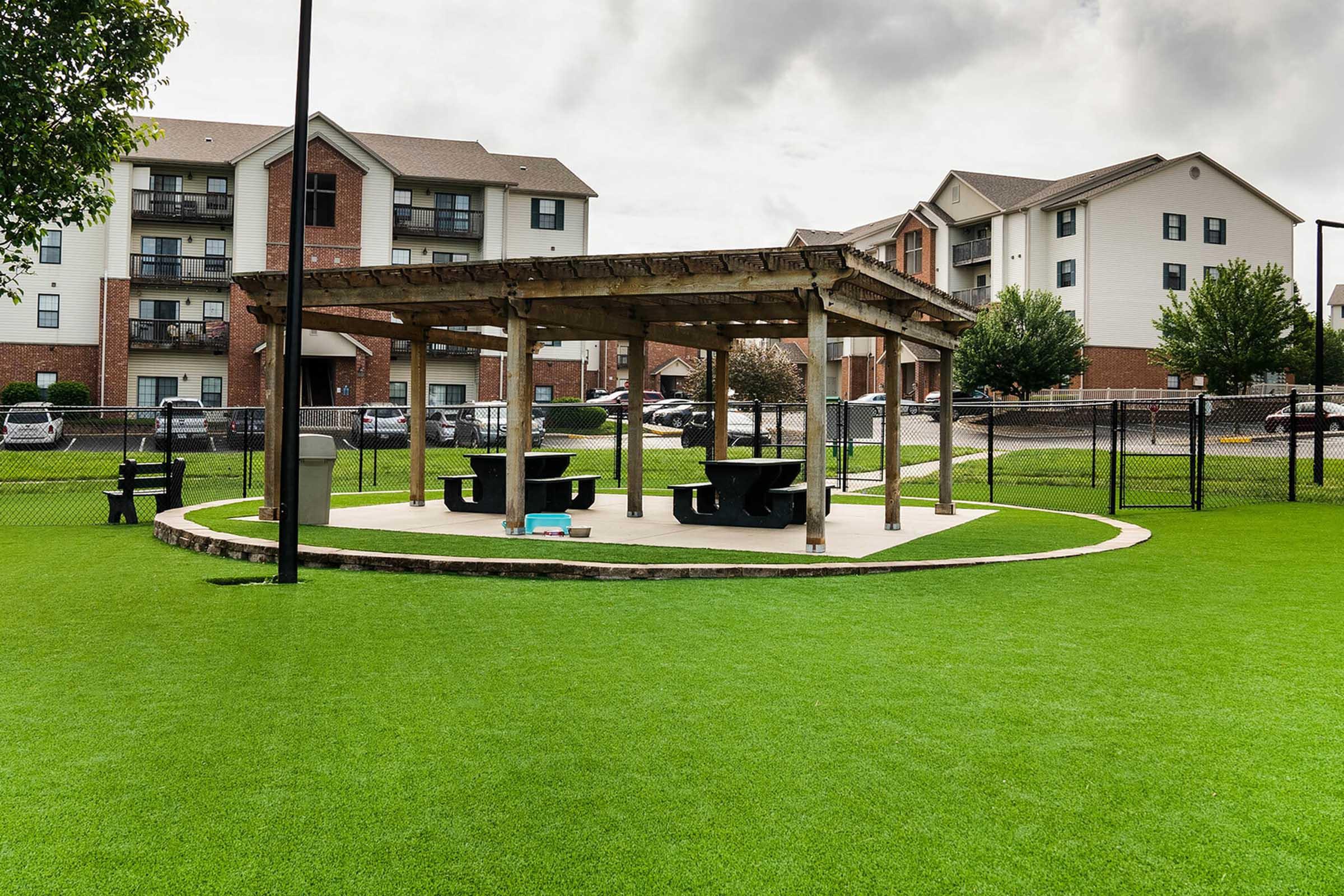 A shaded picnic area with a wooden pergola and black picnic tables, surrounded by a green lawn. In the background, there are multi-story apartment buildings and a parking area. The sky is partly cloudy, creating a pleasant outdoor setting.