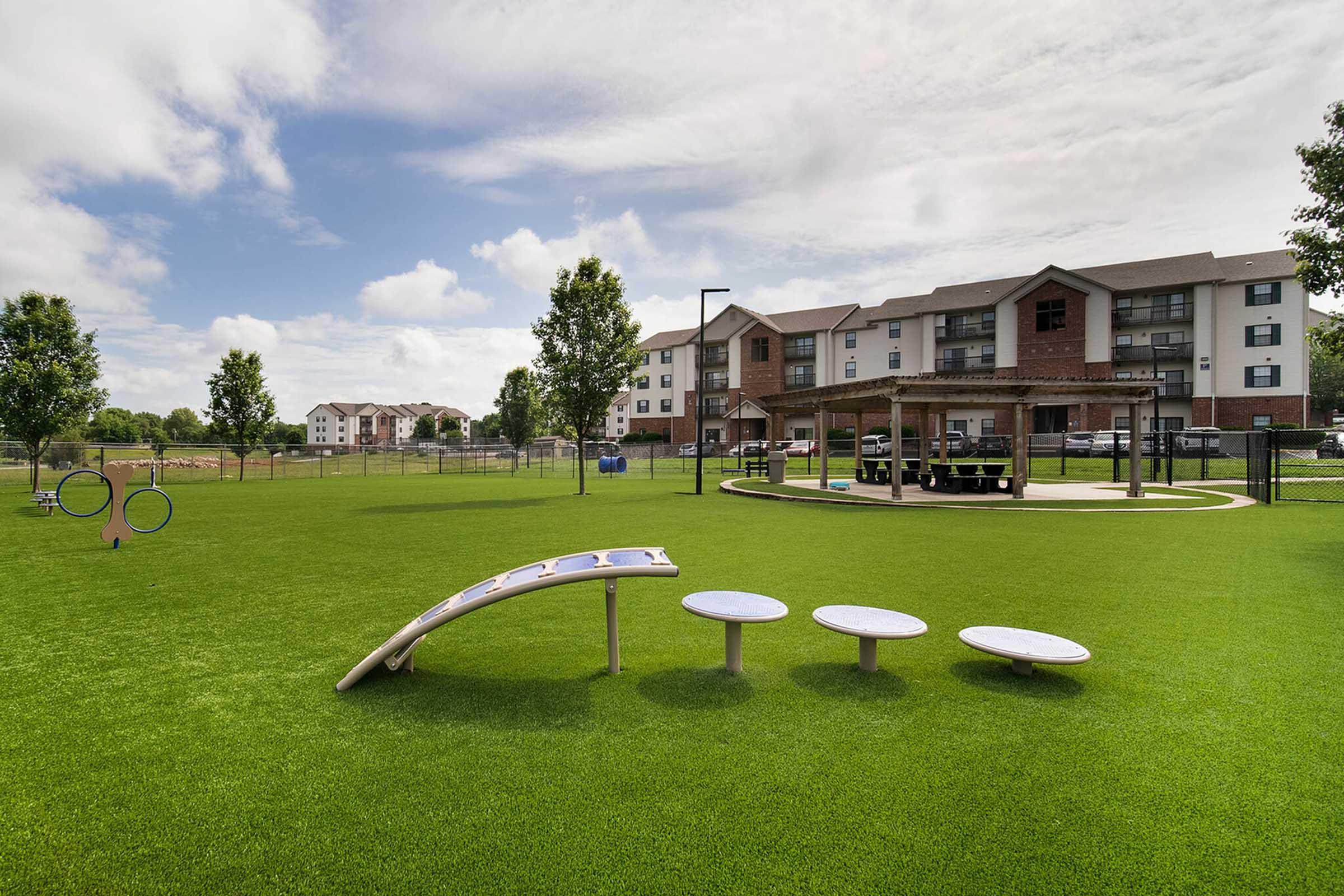 A well-maintained outdoor play area featuring artificial grass, two play structures, and a picnic table under a covered pavilion. In the background, several tan apartment buildings and a blue sky with scattered clouds are visible, creating a serene and inviting atmosphere.