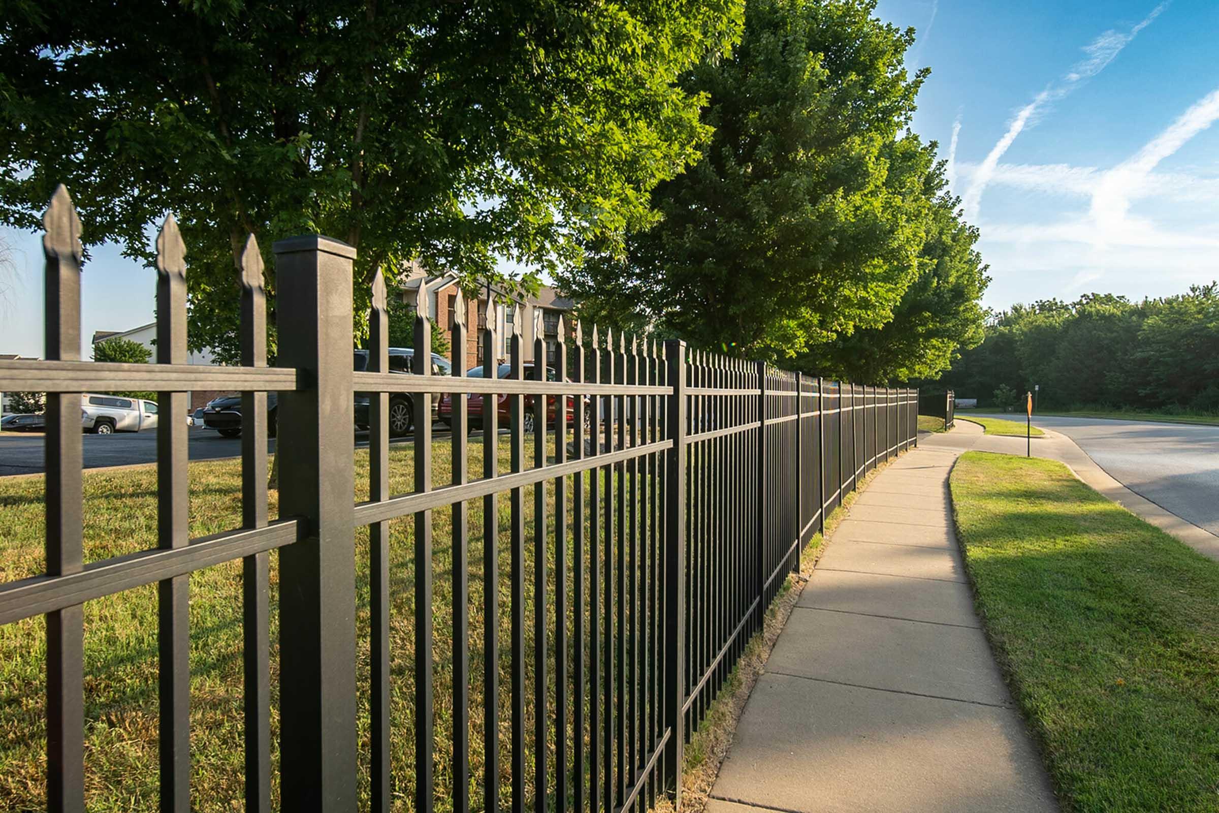 Black ornamental metal fence along a sidewalk, bordered by green trees and grass. The sun is shining, casting a warm glow, with a clear blue sky and wispy clouds in the background. The scene depicts a peaceful suburban area, with vehicles visible in the distance.