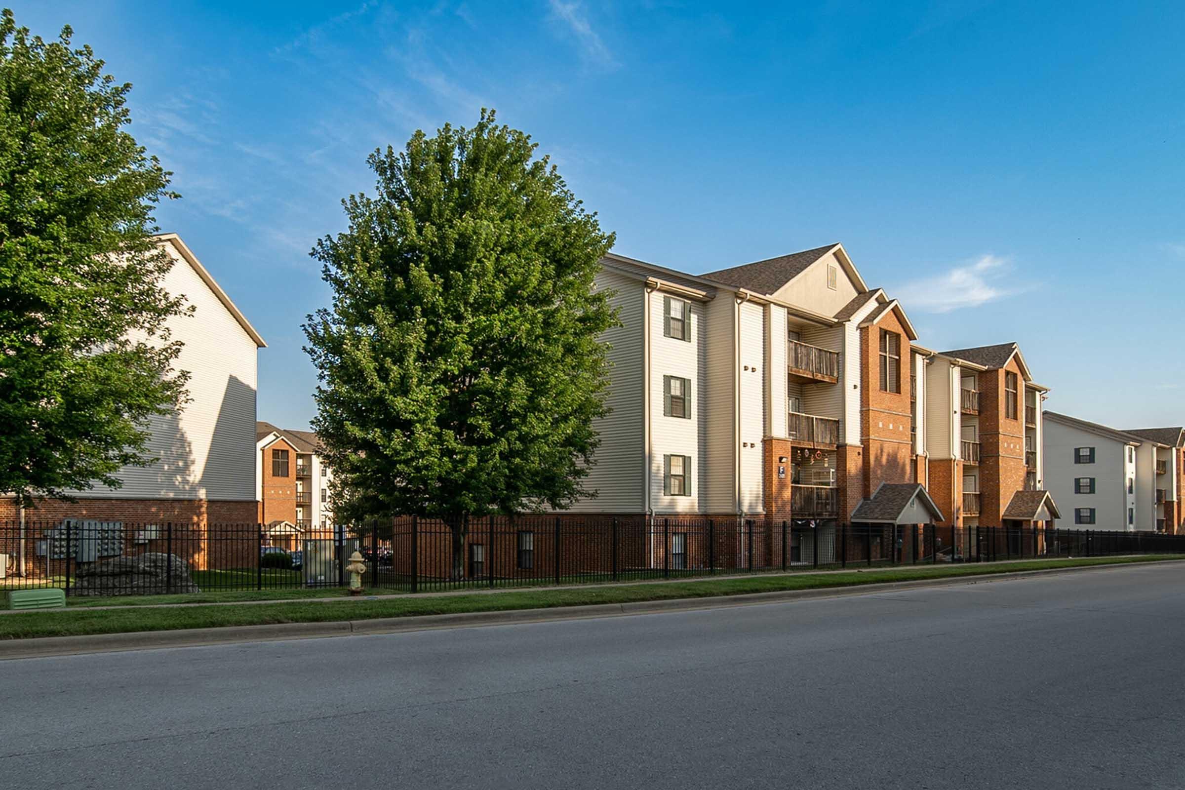 A row of modern apartment buildings with a mix of white and brown exteriors, surrounded by green trees and a well-maintained lawn. The sky is clear and blue, indicating a pleasant day. A fenced area runs along the sidewalk, enhancing the residential atmosphere.