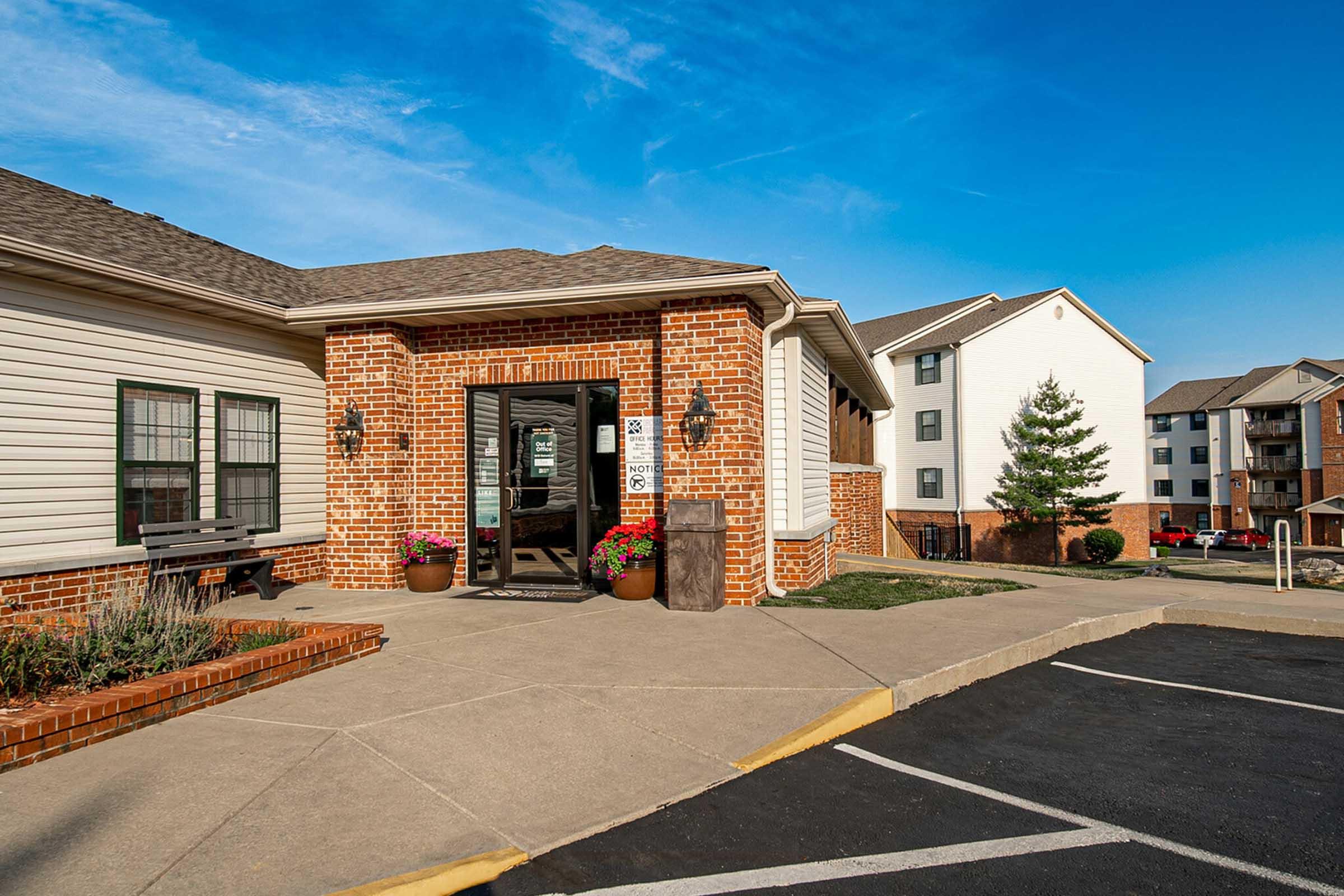 Exterior view of a brick building with a welcoming entrance. The doorway features a glass door and is flanked by colorful flower pots. Surrounding the entrance are landscaped areas with grass and small plants. Clear blue sky and additional buildings visible in the background.