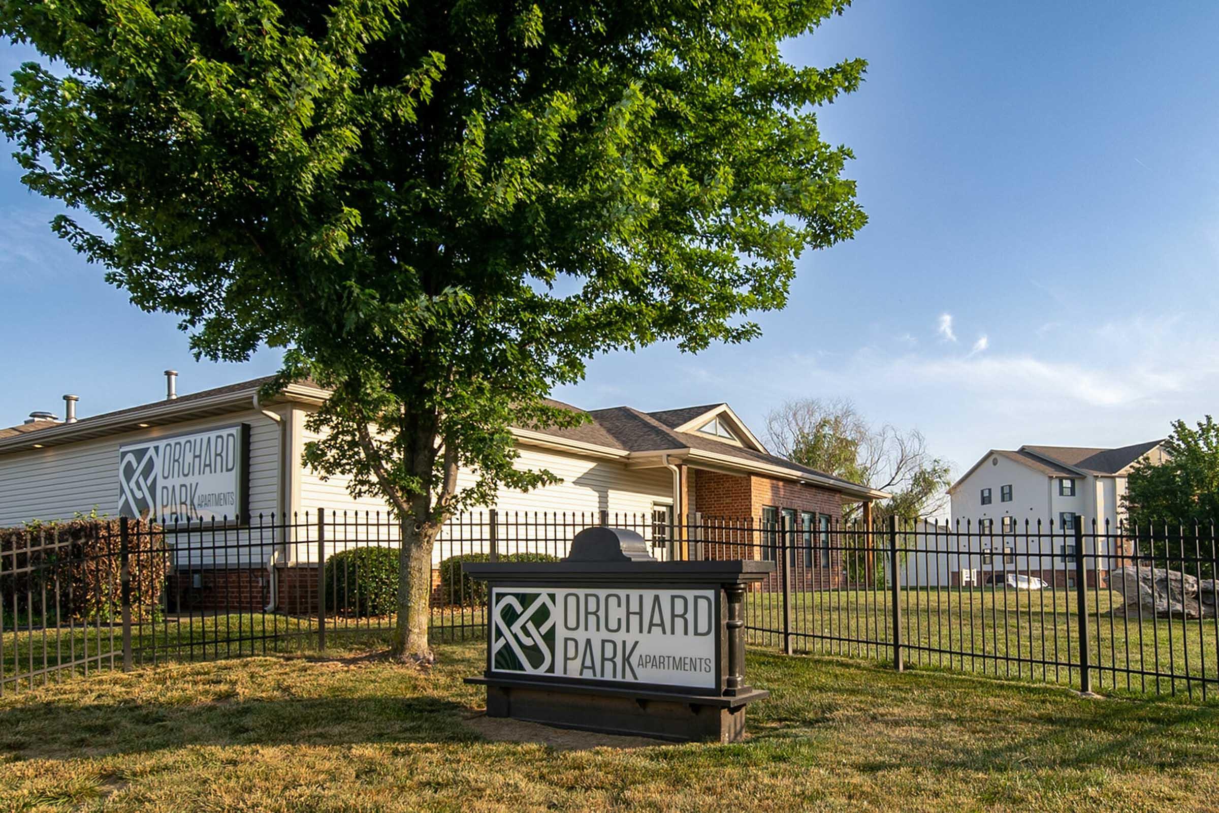A grassy area with a sign for Orchard Park Apartments, featuring a logo and the name prominently displayed. In the background, there are residential buildings and a tree nearby. The scene is bathed in sunlight, suggesting a clear, pleasant day. A fence outlines the property.