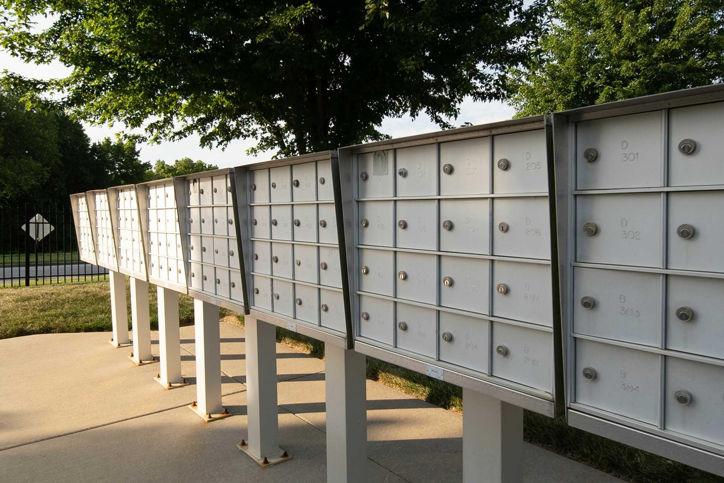 A row of white mailboxes mounted on metal stands, set against a backdrop of green trees and grass. The mailboxes have individual compartments with numeric labels, and the scene is bathed in natural sunlight, suggesting a suburban or residential area.