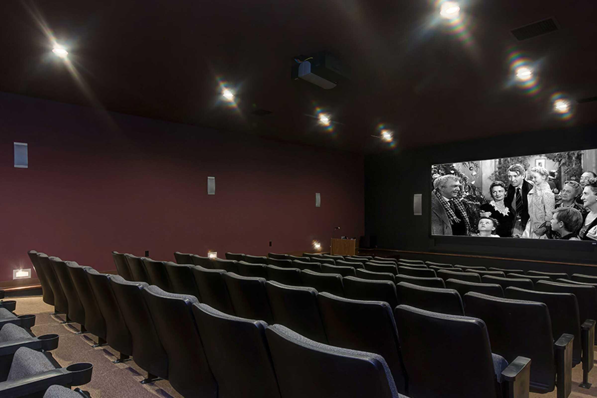 A dimly lit movie theater featuring rows of black seats facing a large screen displaying a black and white film scene. The theater has small wall lights and a projector visible on the ceiling. The ambiance suggests a cozy and intimate viewing experience.