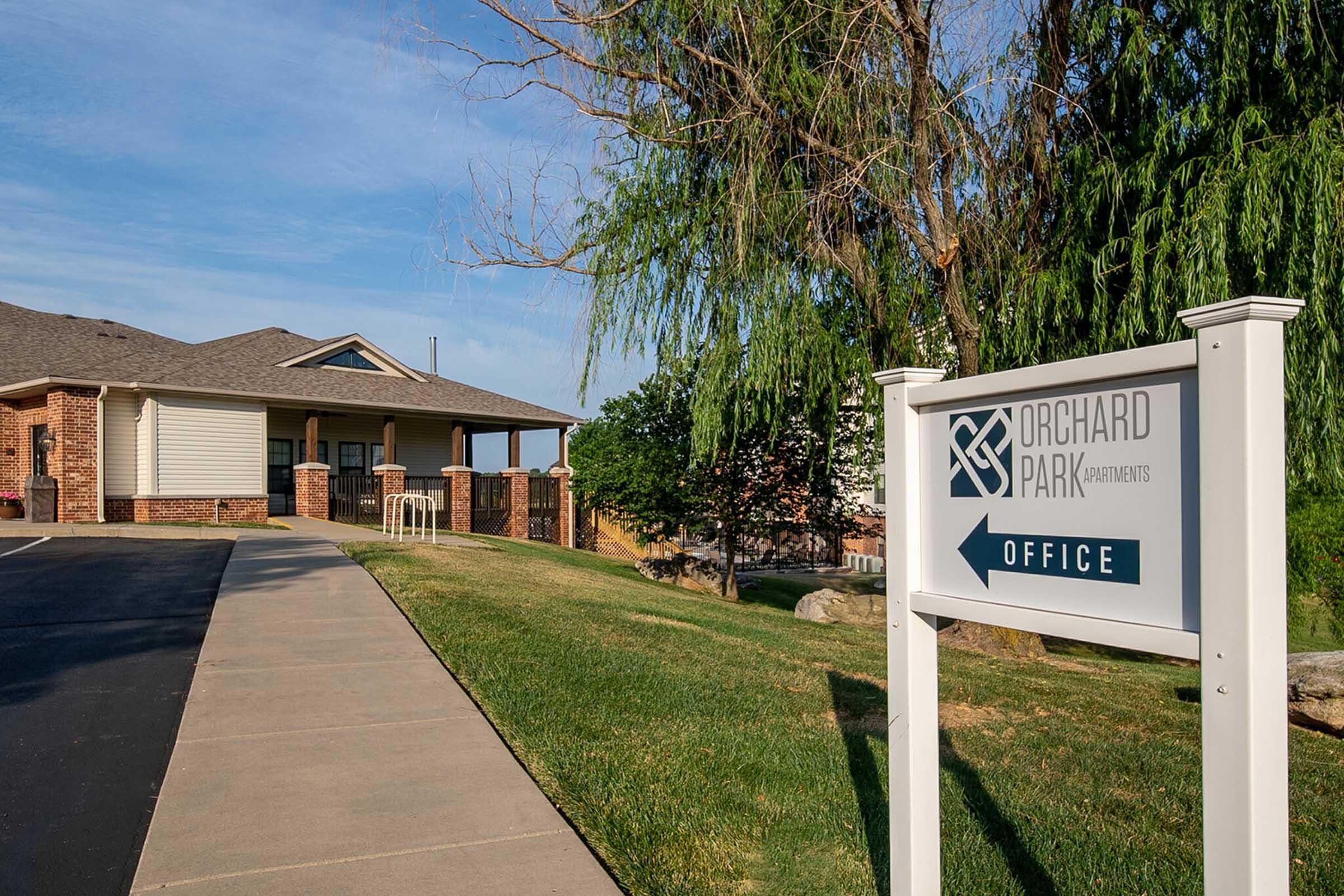 Sign for Orchard Park Apartments Office, with a well-maintained pathway leading to the building. The office features a combination of brick and siding, surrounded by green grass and trees under a clear blue sky.