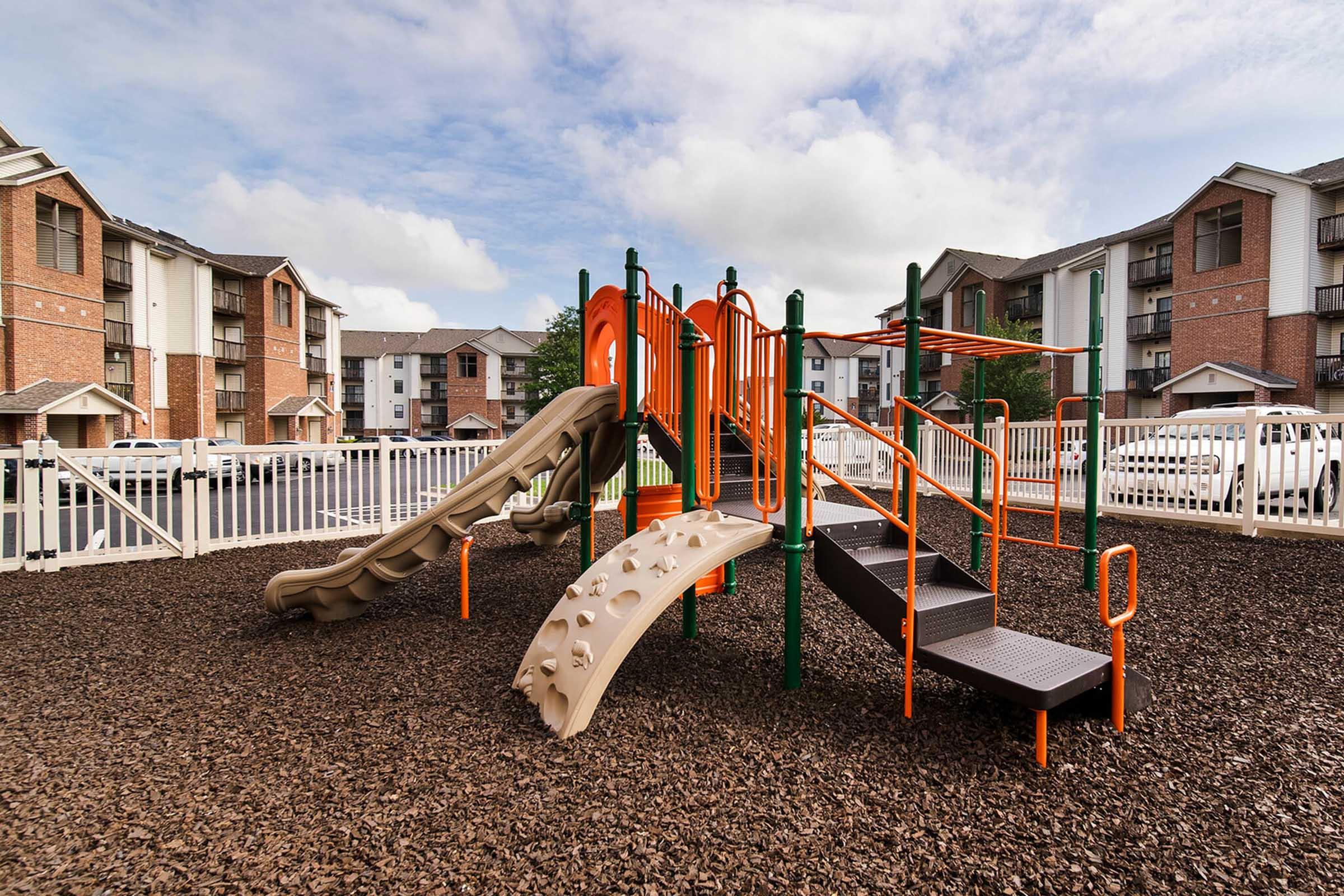 Playground equipment featuring a slide, climbing structures, and stairs, situated on mulch ground. The background shows a fenced area with several multi-story apartment buildings and a cloudy sky.