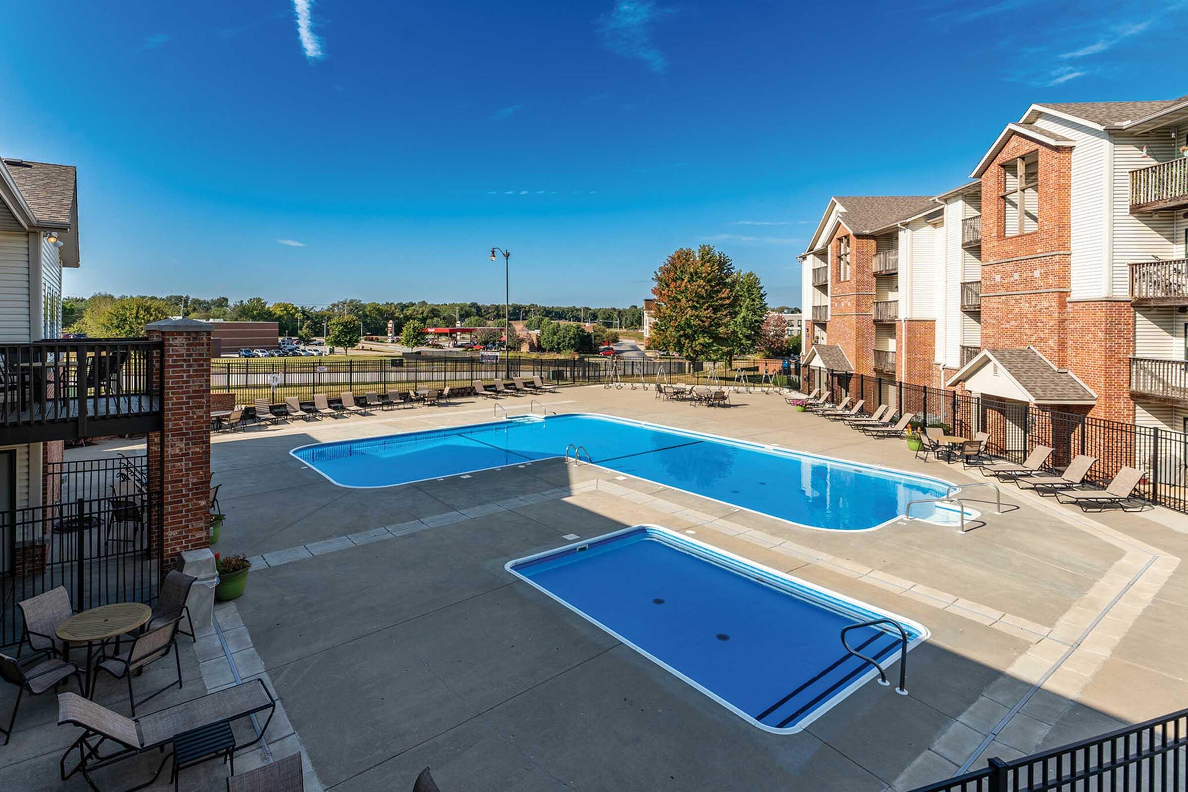 A spacious outdoor swimming pool area surrounded by lounge chairs, with a separate shallow pool for children. The scene features landscaped greenery in the background and several apartment buildings nearby. Clear blue skies above provide a bright and inviting atmosphere.