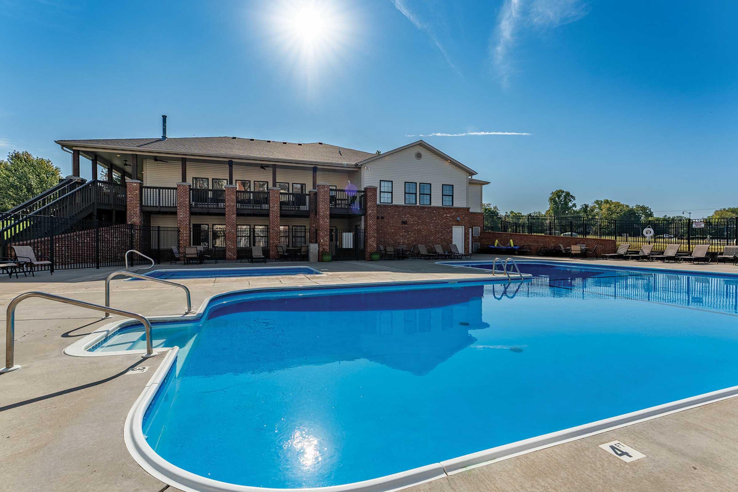 A sunny outdoor pool area featuring a large blue swimming pool with lounge chairs nearby. In the background, a two-story building with balconies is visible, surrounded by green trees and a clear blue sky. The scene evokes a relaxing atmosphere ideal for summer enjoyment.