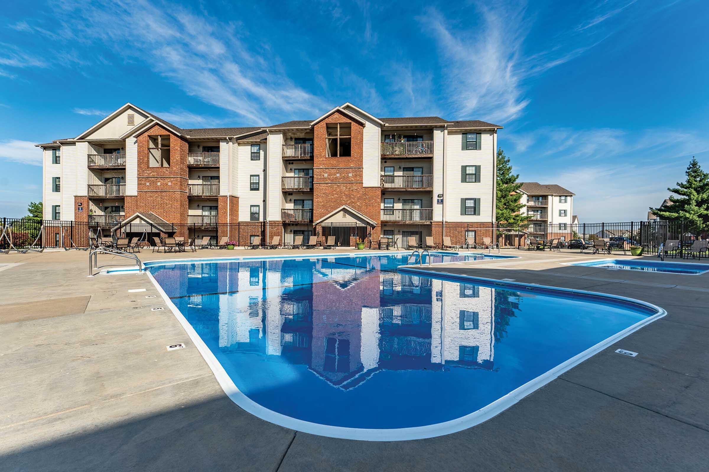 A large swimming pool with a curved shape in the foreground, surrounded by paved decking and lounge chairs. In the background, a modern three-story apartment building with a mix of brick and white siding, and blue skies with a few clouds above.