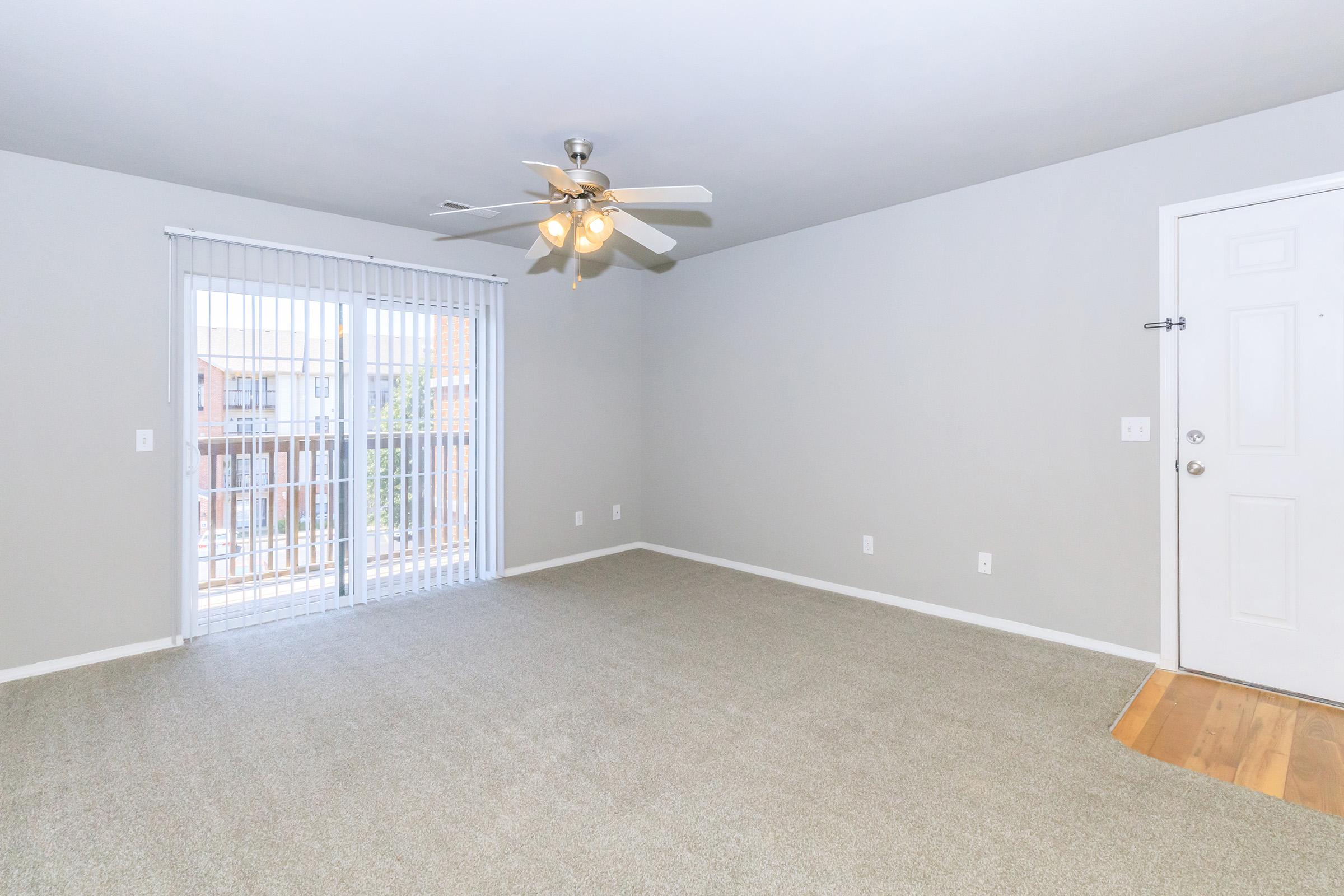 A spacious, well-lit living room featuring light-colored walls, a ceiling fan, and neutral carpet. A sliding glass door leads to a balcony, while the entrance door is visible on the right. The room offers a blank canvas for furniture arrangement and decor.