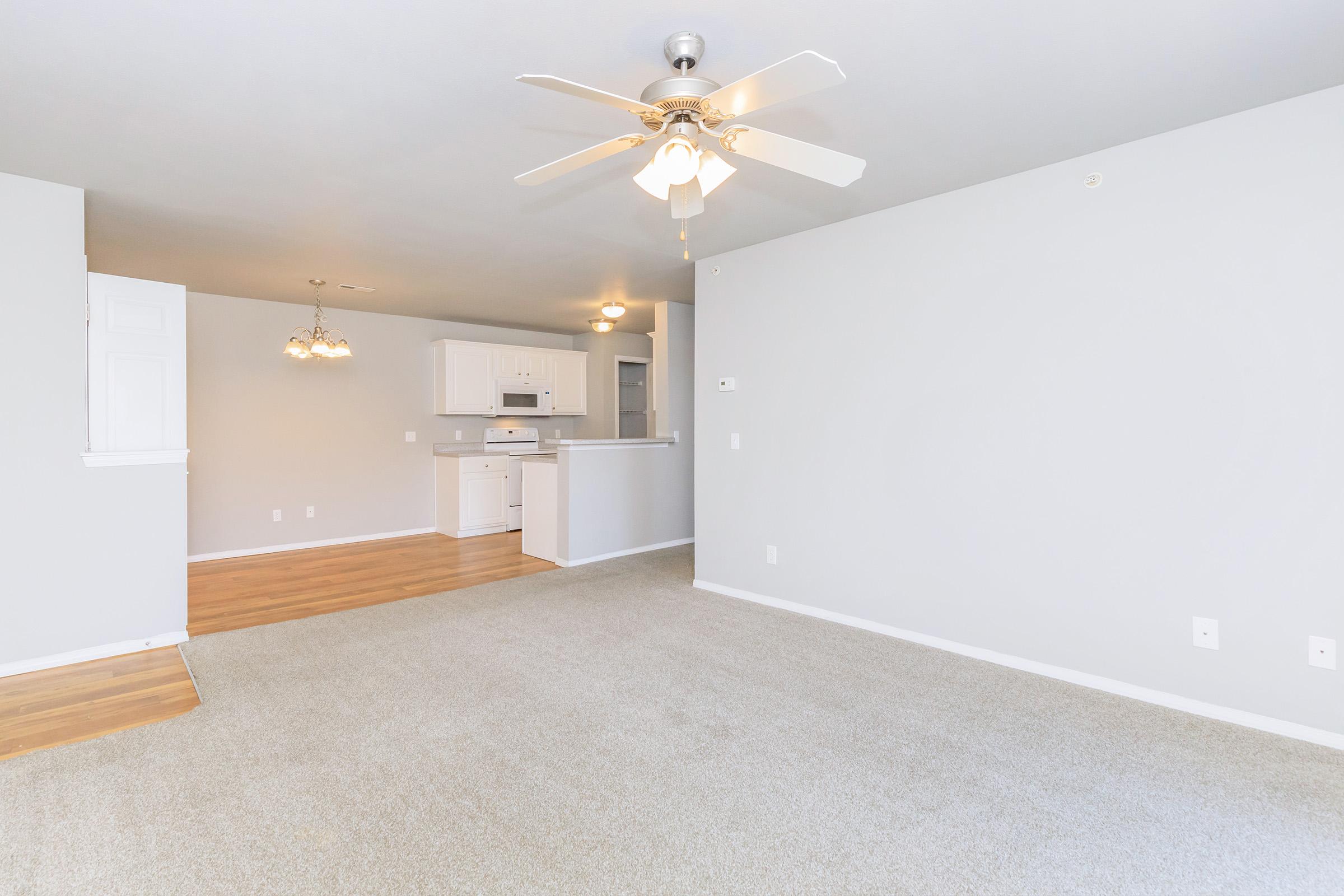 A spacious, light-colored living room with beige carpet and a ceiling fan. The room features a doorway leading to a kitchen area with white cabinets and appliances. Two light fixtures hang above, and natural light brightens the space. The walls are painted in soft gray tones.