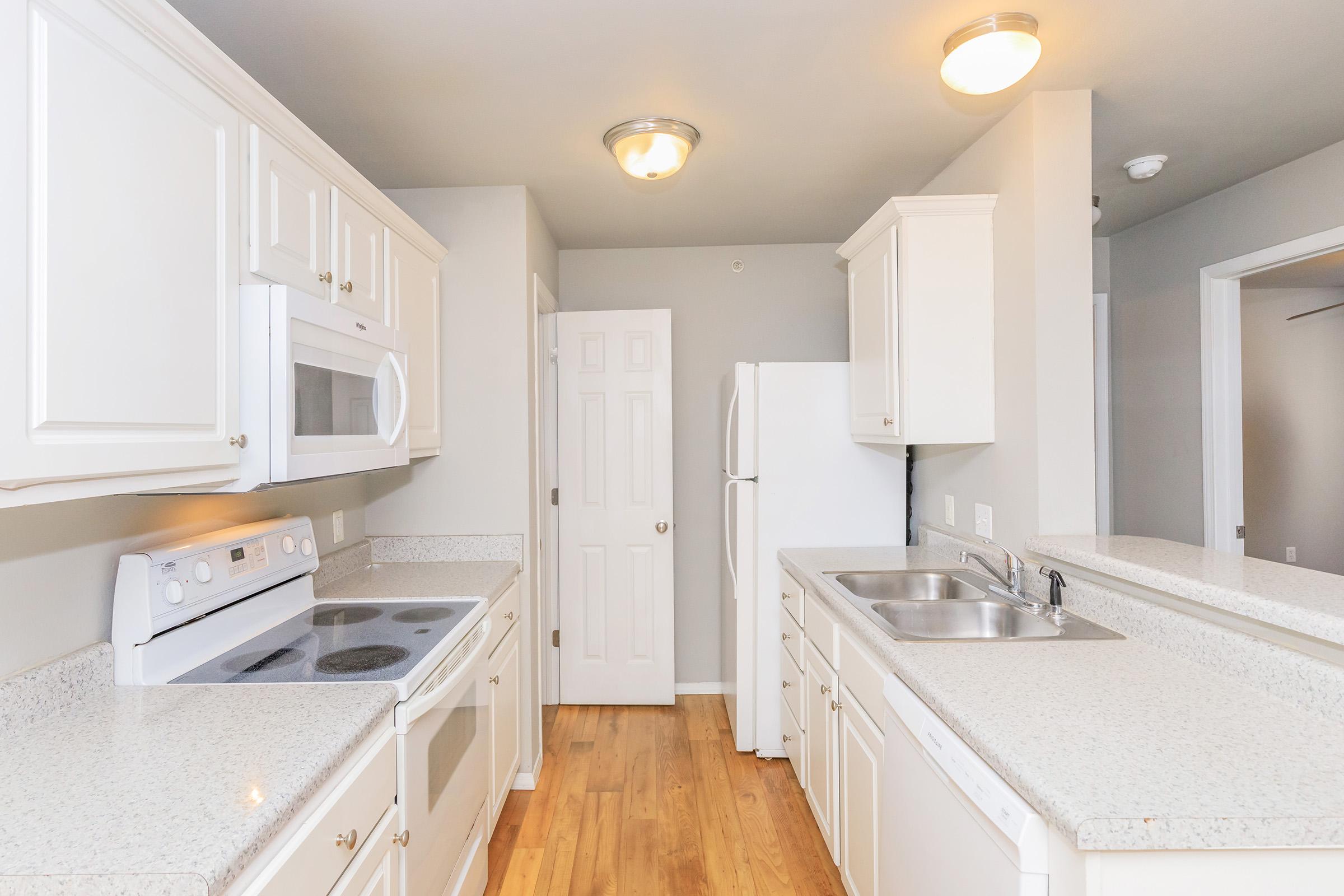 A modern kitchen featuring white cabinetry, granite countertops, a white stove, microwave, and refrigerator. The open layout includes a double sink, and light-colored walls with wood flooring. A closed door leads to another room, adding to the spacious feel. Bright lighting enhances the inviting atmosphere.
