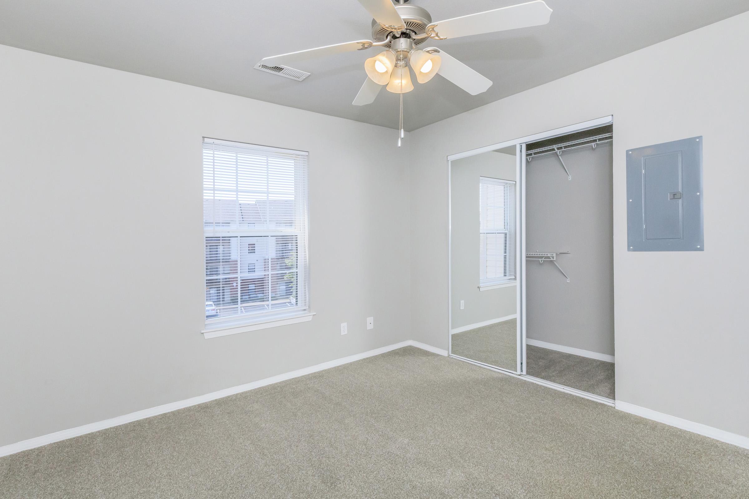 A sparsely furnished room with light gray walls and carpet. It features a ceiling fan with light, a window with blinds, and a mirrored closet door. The space is well-lit and has a clean, neutral atmosphere, ideal for various decor styles.