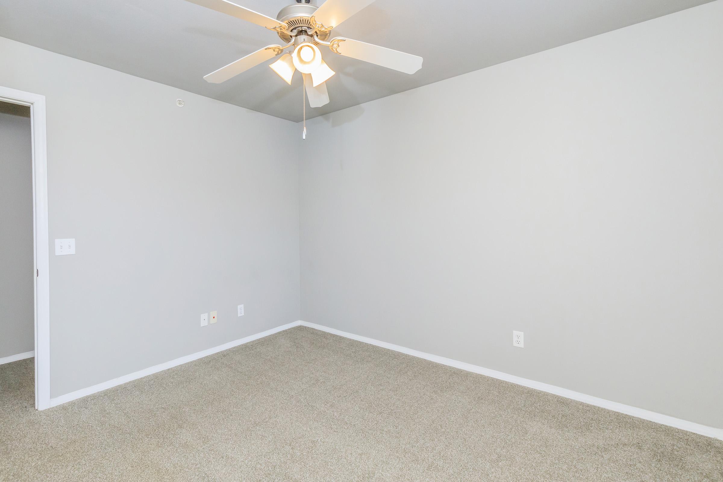 An empty room with light gray walls and a ceiling fan. The floor is covered with beige carpet, and there is a white door on the left side. The room is well-lit, featuring natural light and a spacious feel with no furniture present.