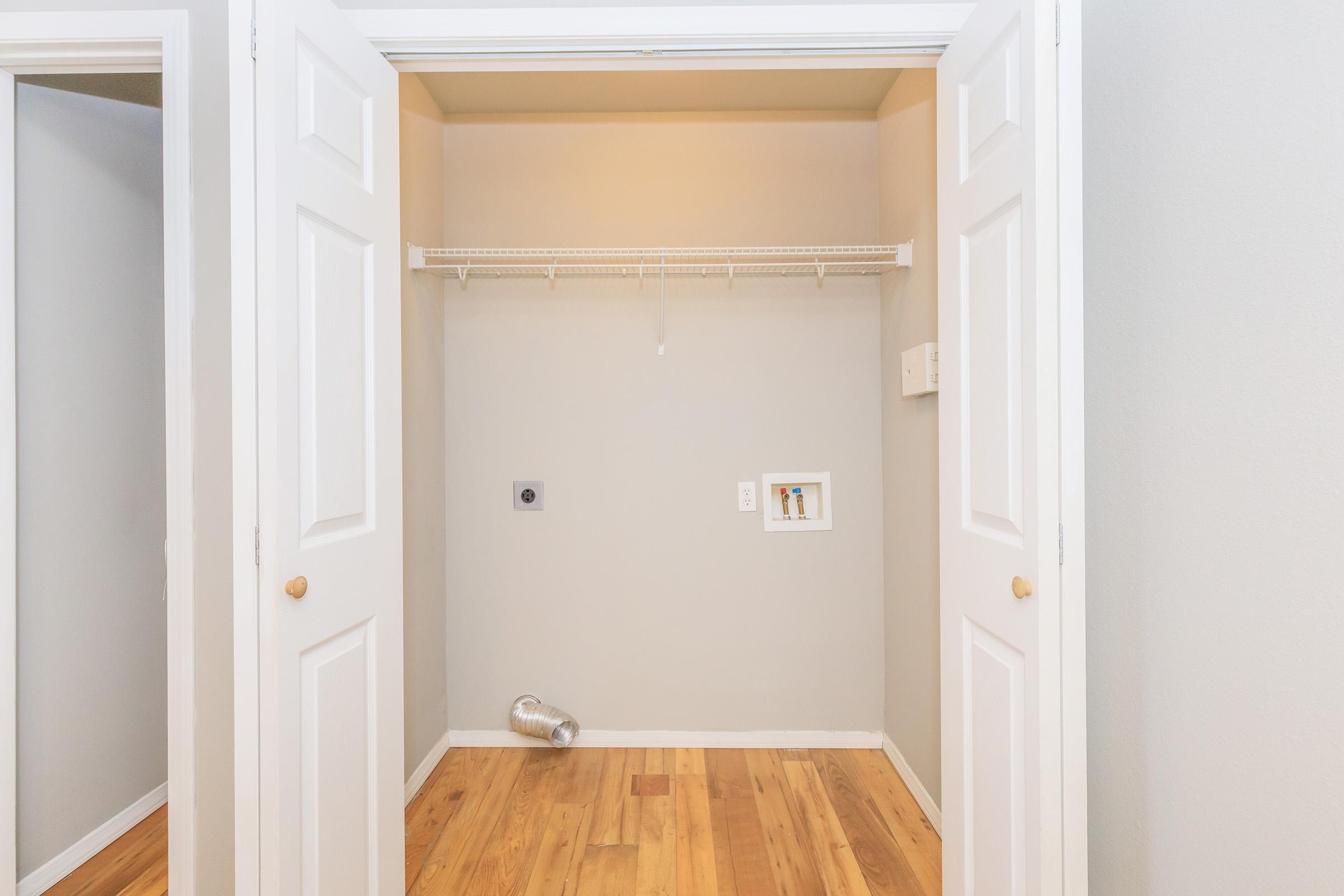 A small, empty closet with double doors open. The walls are painted light gray, and there is a hanging wire shelf inside. The floor is wooden, and there is a small metallic vent or dryer hose on the floor. A power outlet is visible on the side wall, along with a wall-mounted cable box.