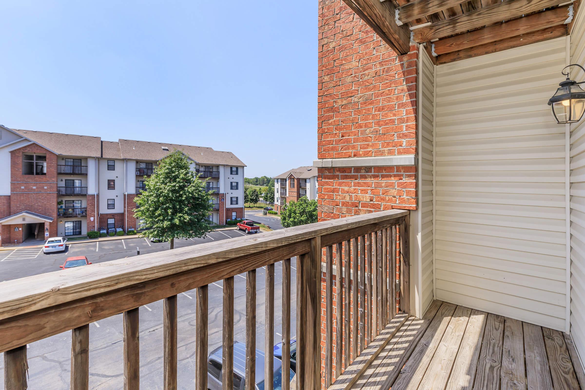 View from a balcony overlooking a parking lot surrounded by residential buildings. The balcony features wooden flooring and a brick exterior, with a lamp hanging on the side. Bright sunny day with clear blue skies.