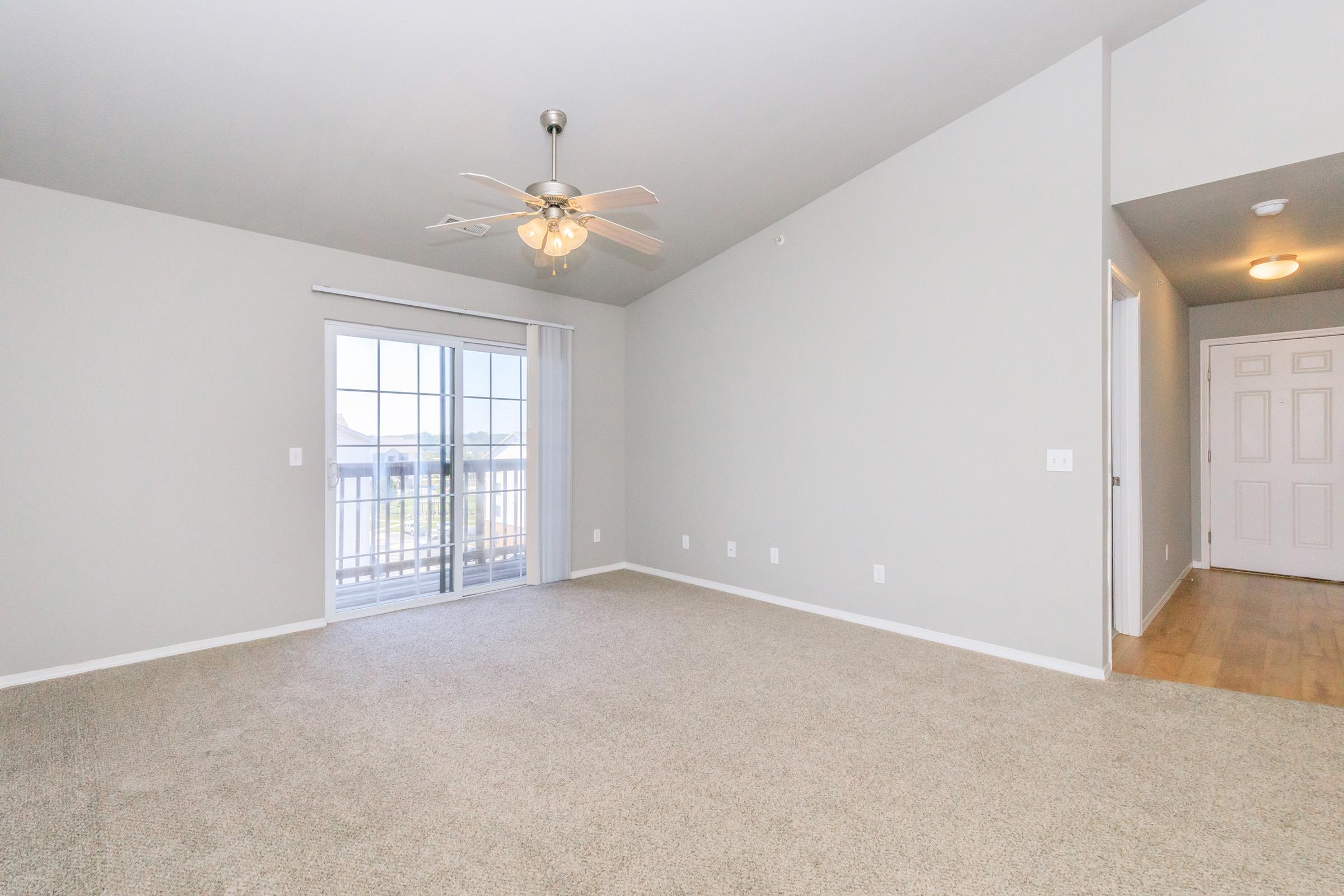 Spacious, empty room featuring light beige carpet, a ceiling fan, and off-white walls. A sliding glass door leads to a balcony, allowing natural light. The room has no furniture, emphasizing its openness. A doorway is visible on the right, suggesting access to another area.