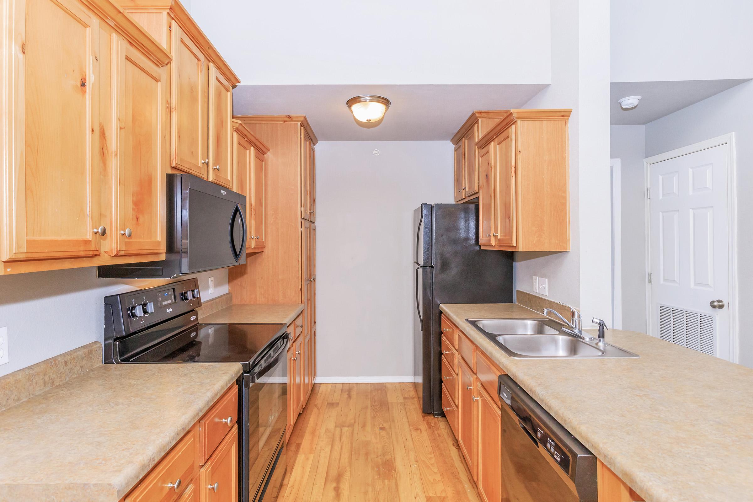 A modern kitchen featuring wooden cabinetry, stainless steel appliances including a microwave and oven, a black refrigerator, and a double sink. The countertops are beige, and the flooring is wooden. The walls are painted light gray, and there is a door leading to another area.