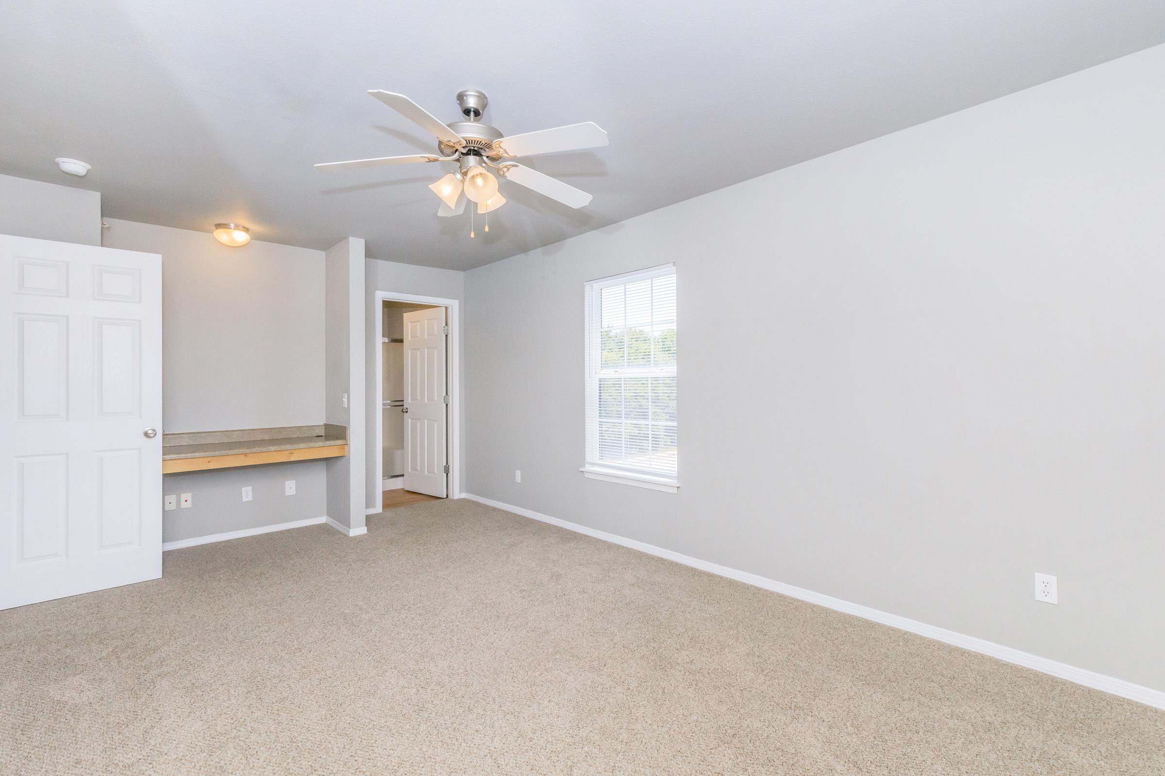 A spacious, empty room with light gray walls and carpeted flooring. It features a ceiling fan, a window letting in natural light, and a door leading to another area. There's also a small counter surface along one wall, creating a versatile space suitable for various uses.