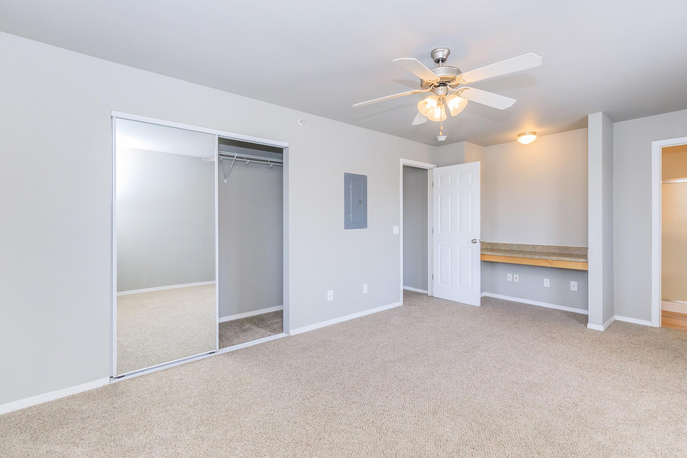 A spacious, well-lit bedroom featuring light gray walls, a ceiling fan, and beige carpet. There's a mirrored closet on the left, a white door leading to another room, and a small brown shelf along the wall. Bright natural light enters from the right side, enhancing the airy feel of the space.