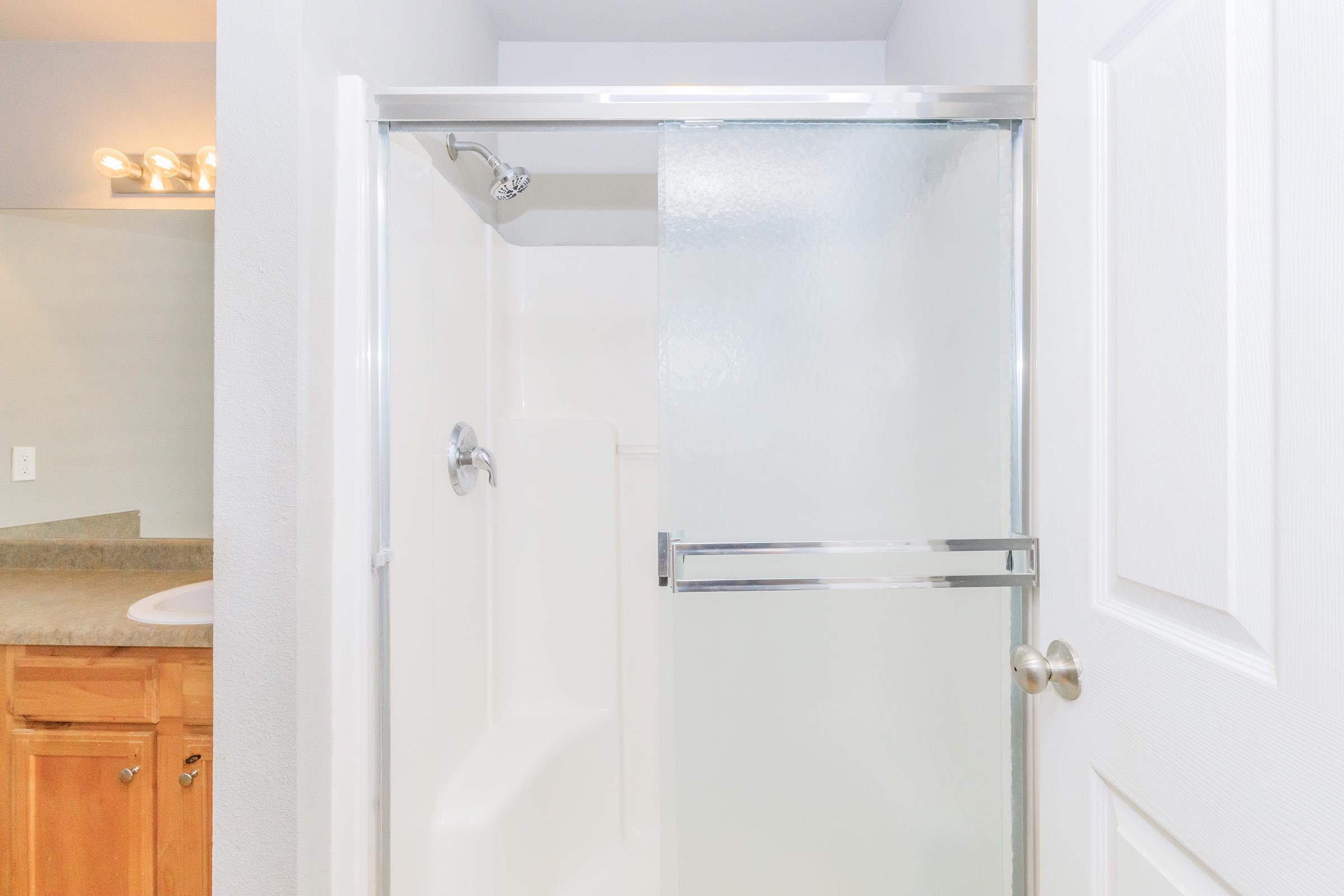 A clean, modern bathroom featuring a glass shower enclosure with a sliding door. The walls are painted light, and there's a countertop and mirror visible in the background. The space is well-lit with a row of vanity lights above the mirror.